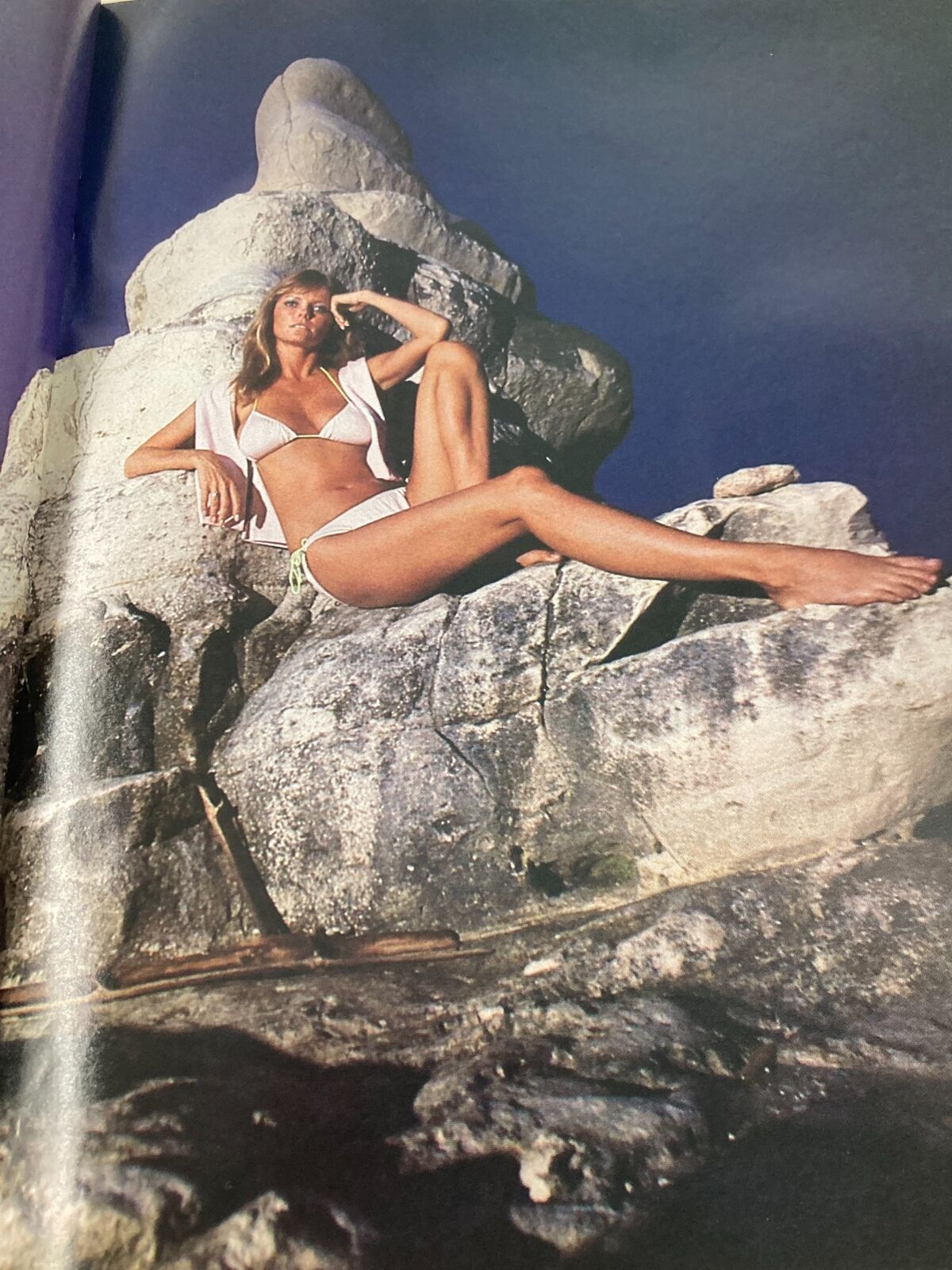 Image from VTG Sports Illustrated Magazine February 14 1983, featuring a stunning model in a bikini, relaxing on a rock formation with a dramatic background, highlighting 1980s beach fashion.