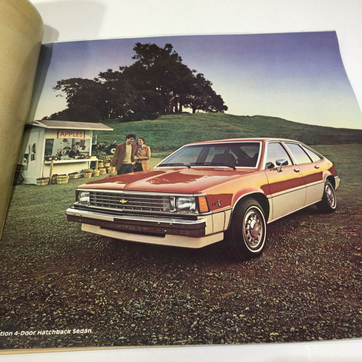 1981 Chevrolet Citation 4-Door Hatchback Sedan Car Sale Brochure image displaying a bright orange Chevrolet Citation parked on a grassy hill with a vintage market setup in the background.