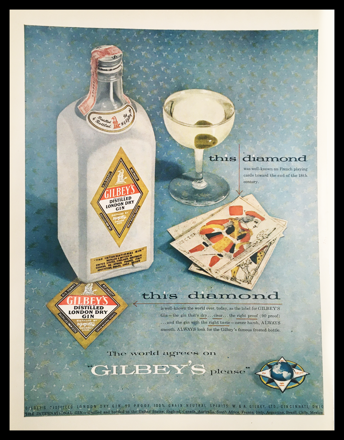 1955 Gilbey's Distilled London Dry Gin Vintage Print Ad showcasing a decorative bottle of gin alongside a cocktail glass and illustrated cards, reflecting mid-20th century design and marketing aesthetics.