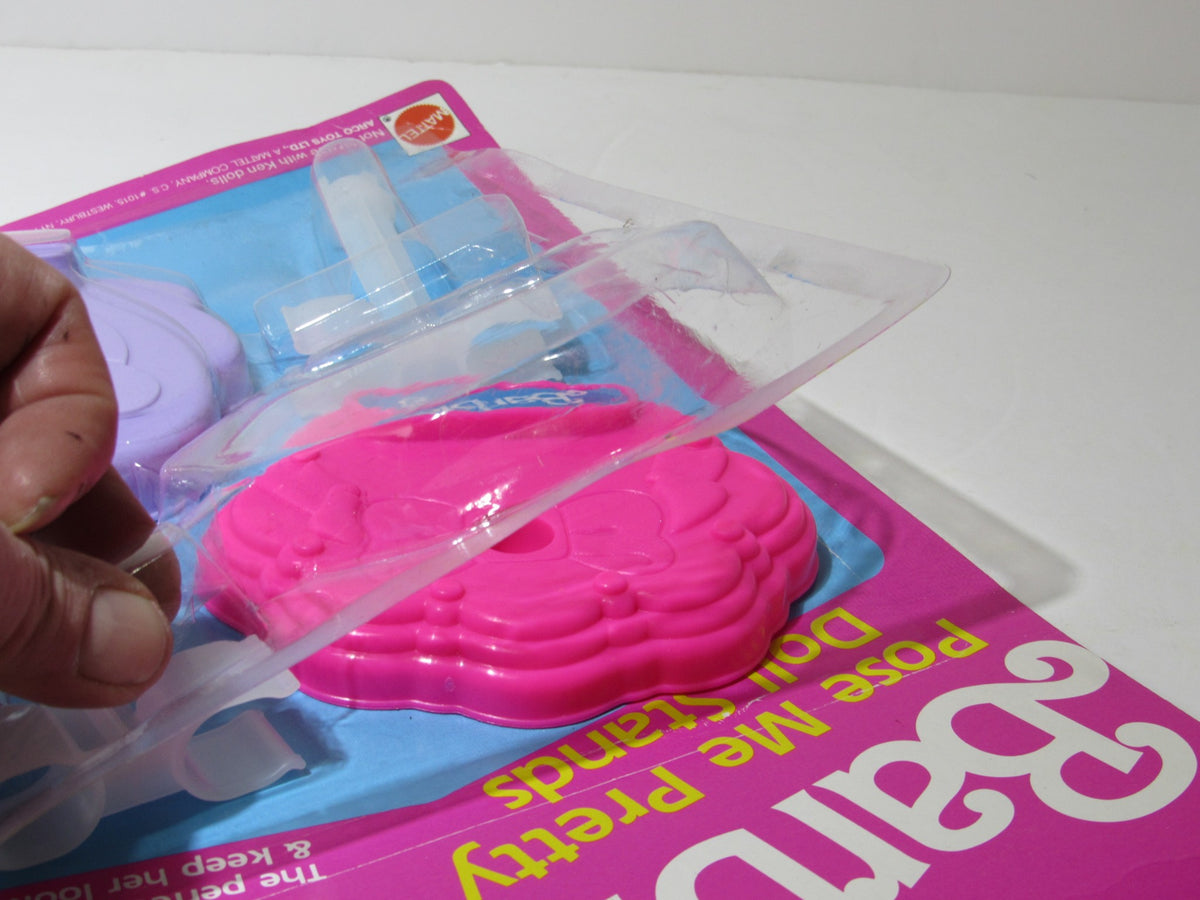 Close-up of the VTG Mattel BARBIE Pose Me Pretty Doll Stands packaging with a hand lifting the plastic cover, revealing the colorful doll stands.