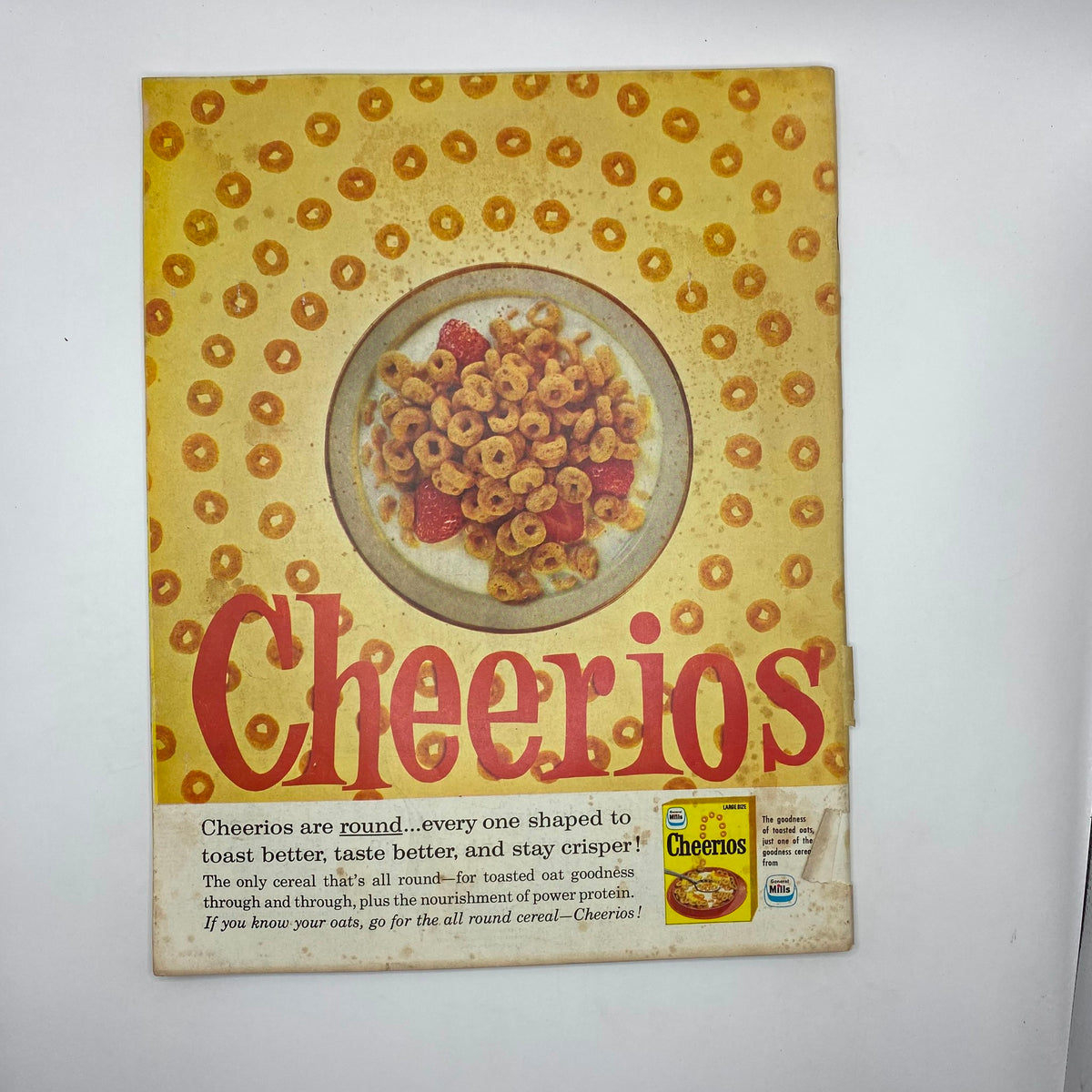 Vintage Cheerios cereal advertisement featuring a colorful bowl of Cheerios with strawberries and a yellow background. The title 'Cheerios' prominently displayed, highlighting its round shapes and crisp taste. The ad promotes Cheerios as the all-round cereal for nourishment and toasty goodness.