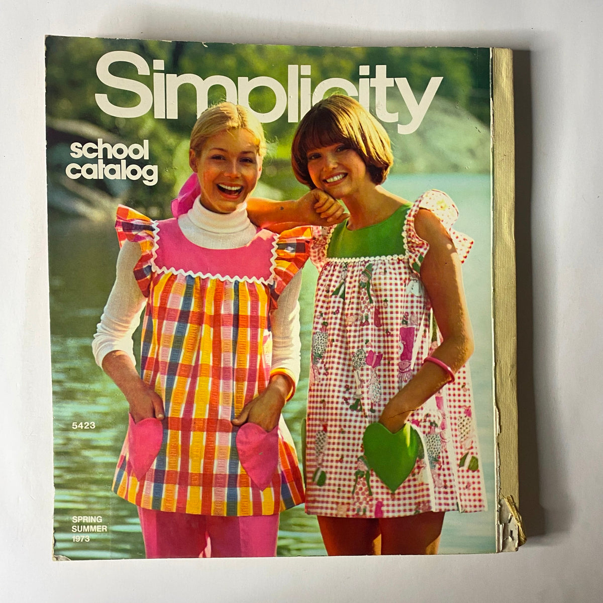 1973 Simplicity Spring Summer School Catalog cover, featuring two young women modeling vibrant, patterned smocks with large pockets, embodying 1970s fashion.