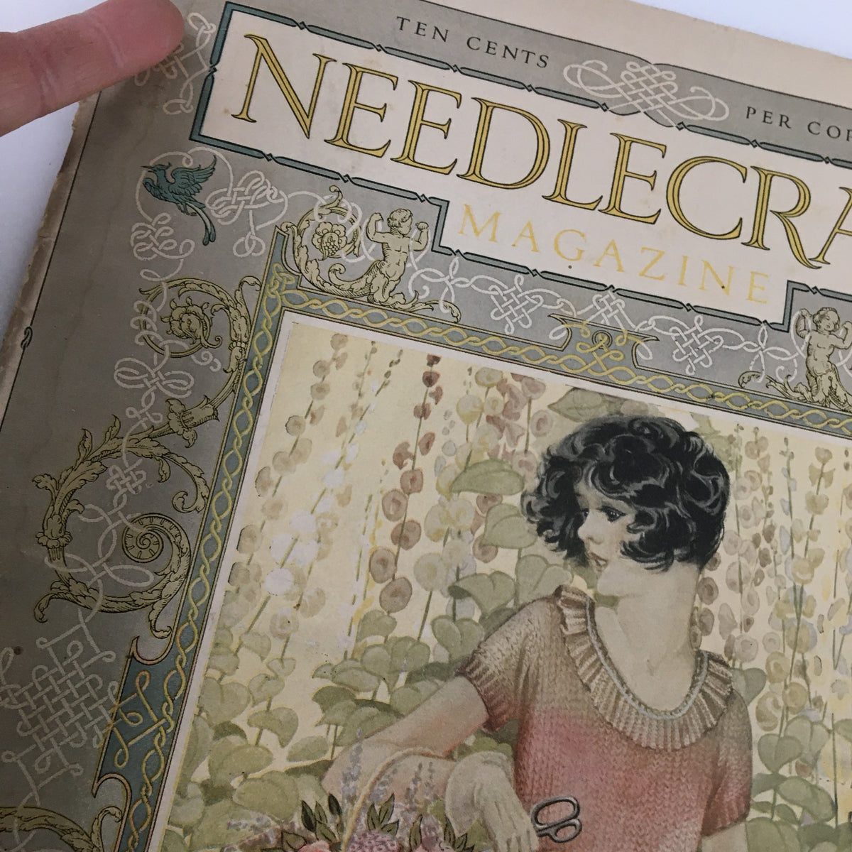 Close-up of the cover of VTG Needlecraft Magazine June 1926 displaying detailed artwork of a woman with a floral basket, highlighting design elements and magazine title.
