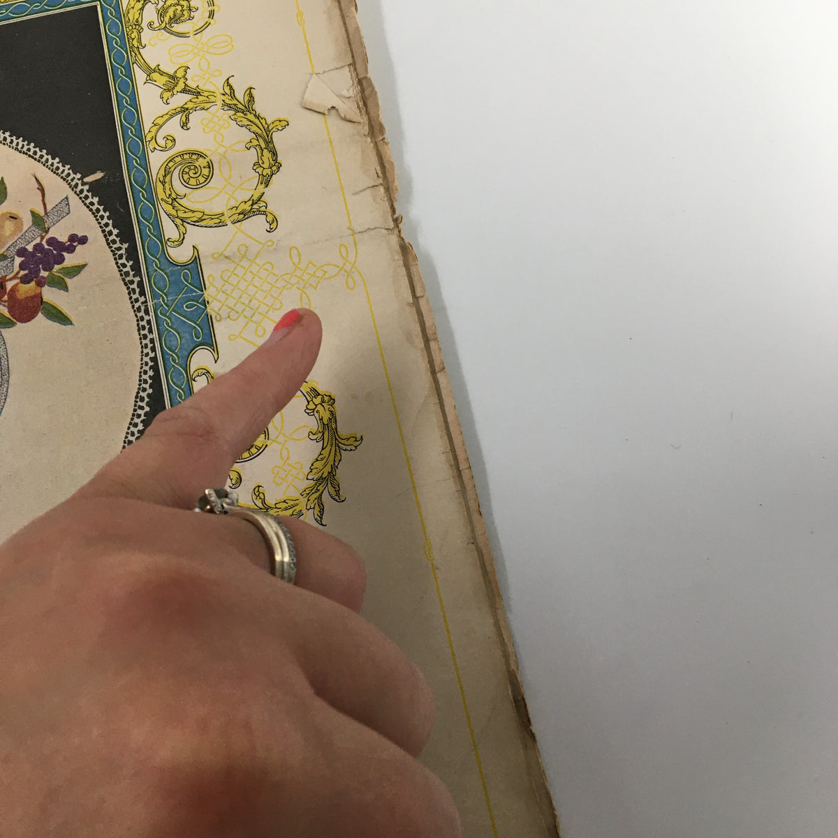 Detailed shot of the edge of VTG Needlecraft Magazine December 1921 with a finger pointing to decorative elements on the cover, highlighting its vintage craftsmanship.