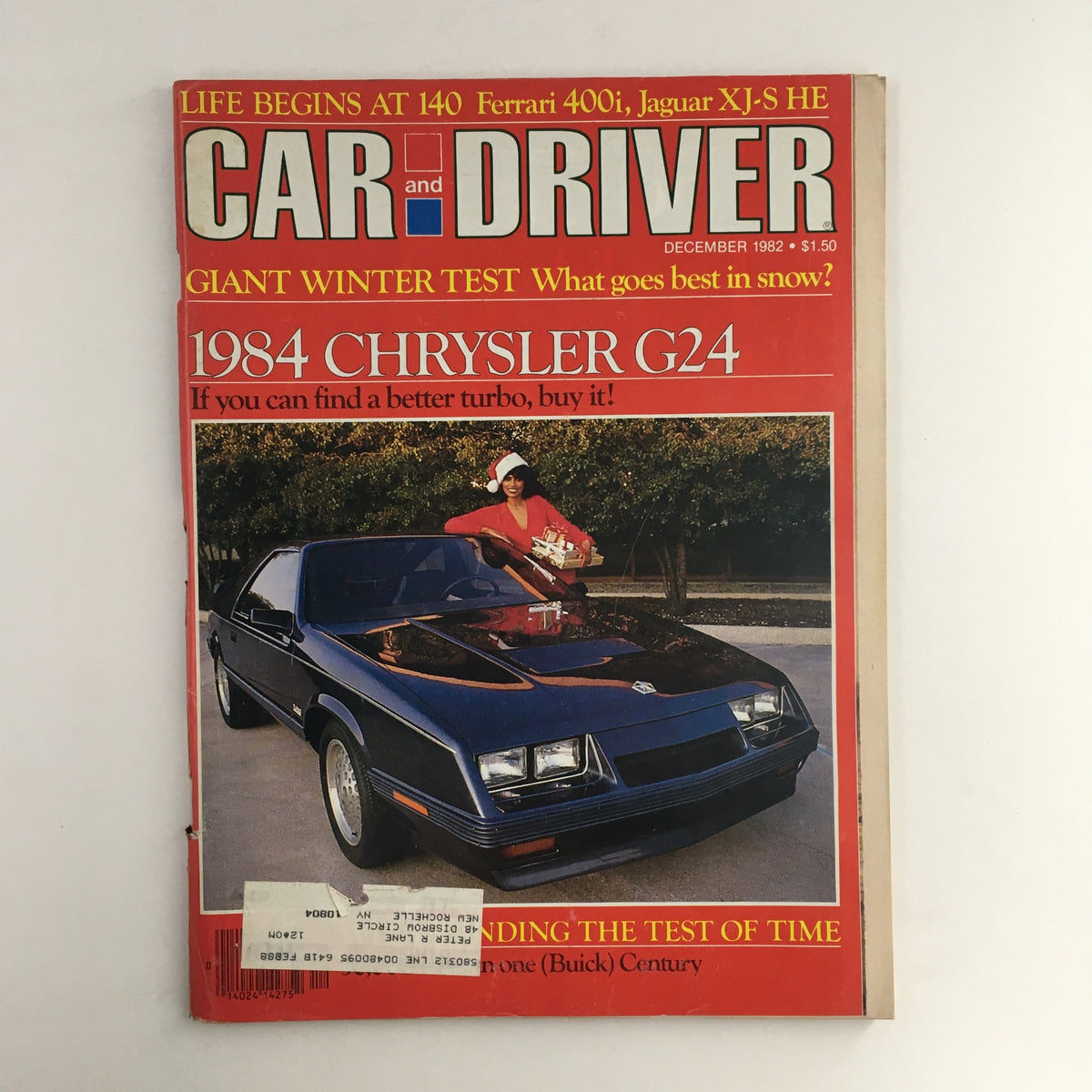 Cover of Car and Driver Magazine December 1982 featuring the Ferrari 400i, Jaguar XJ-S HE, and Chrysler G24, showcasing a woman in holiday attire with a gift beside a Chrysler G24 sports car.