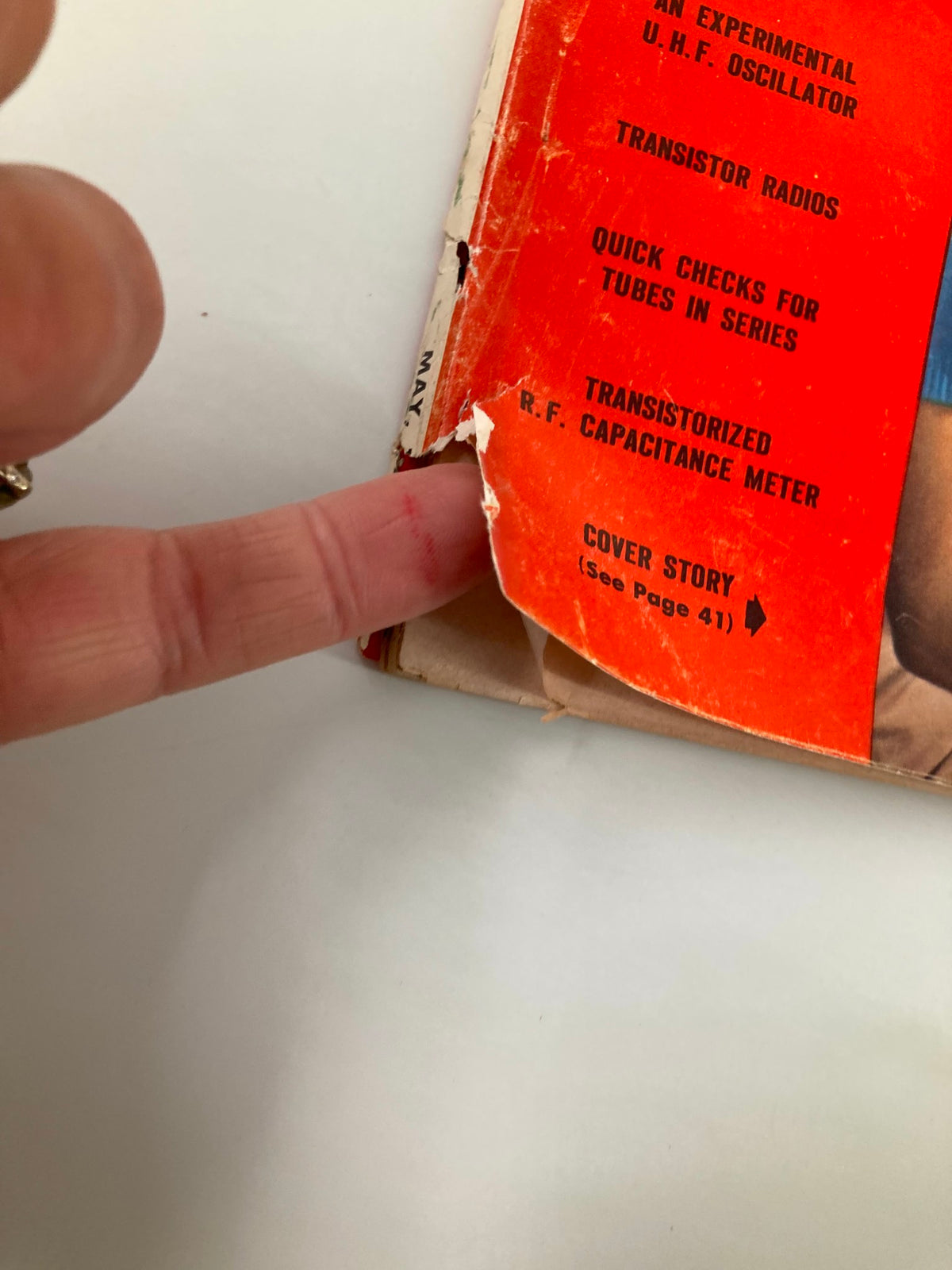 Close-up of the cover of VTG Radio & Television News Magazine May 1956, highlighting wear and tear with a finger pointing at the damaged area, showcasing its vintage character and historical significance.