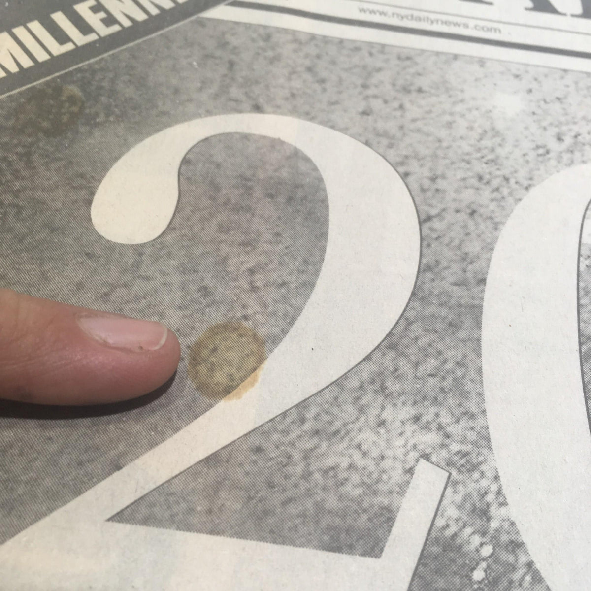 Finger pointing at a stain on the NY Daily News newspaper dated 1/1/2000, highlighting age-related imperfections while celebrating the largest peacetime crowd in Times Square.