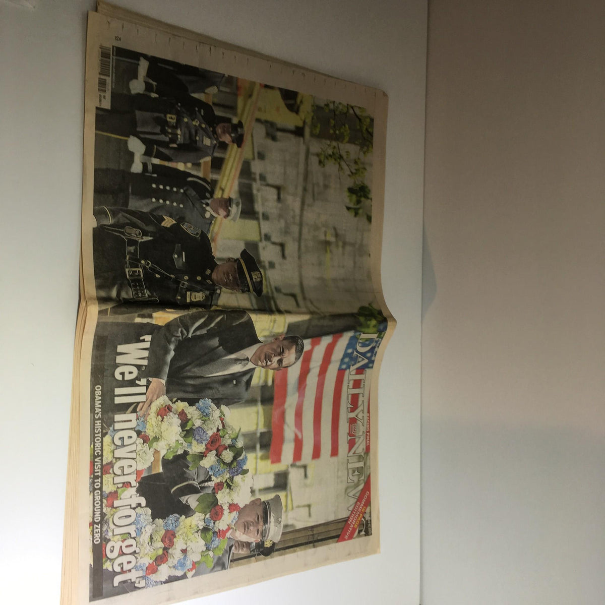 Side view of the NY Daily News cover from May 6, 2011, showcasing Obama's visit to Ground Zero with floral tributes and the phrase 'We'll Never Forget.'