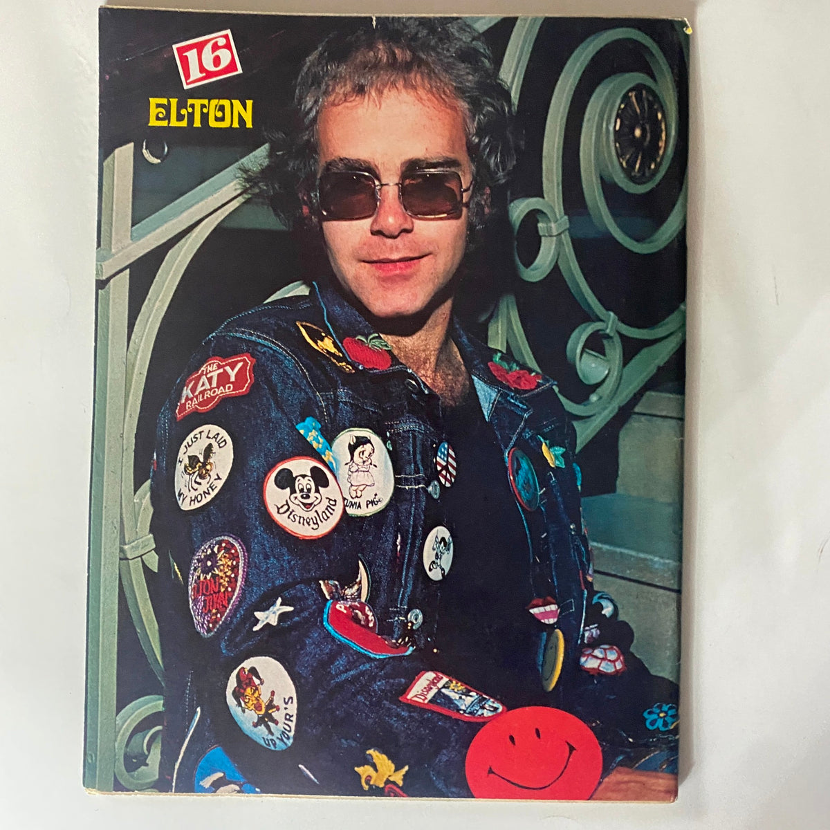Cover of VTG 16 Magazine June 1976 featuring Elton John dressed in a denim jacket adorned with various colorful pins and badges.