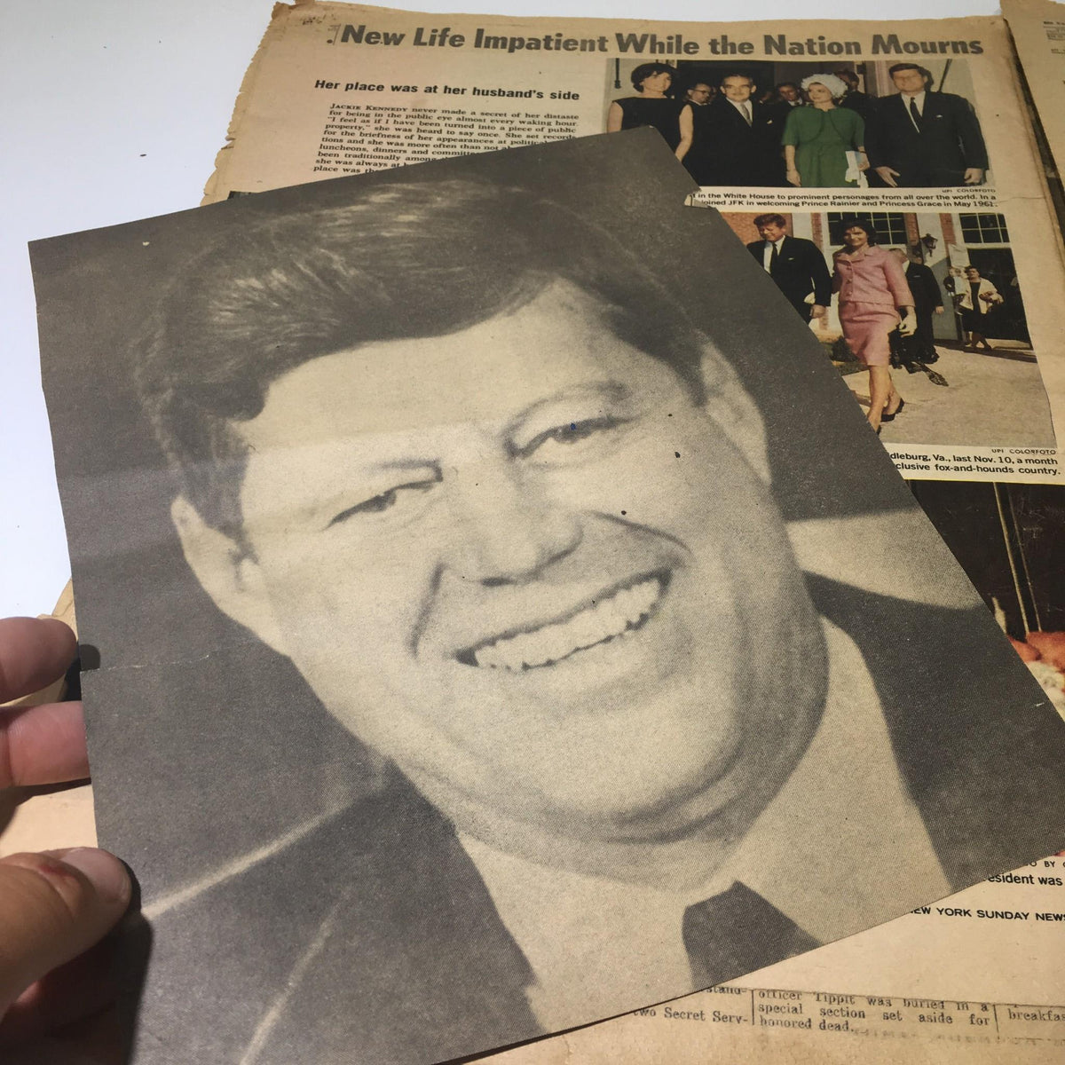 Portrait of John F. Kennedy held alongside a vintage New York Daily News from November 26, 1963, titled 'We Carry On', showcasing a historic moment.
