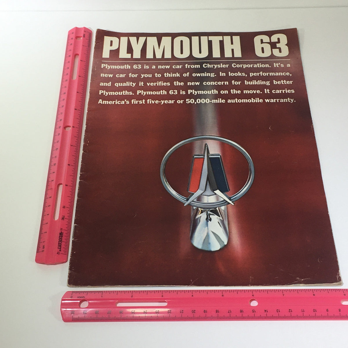 Detailed view of the 1963 Plymouth by Chrysler Corp. brochure cover with a ruler for scale, highlighting the brochure design featuring the Plymouth logo and promotional text.