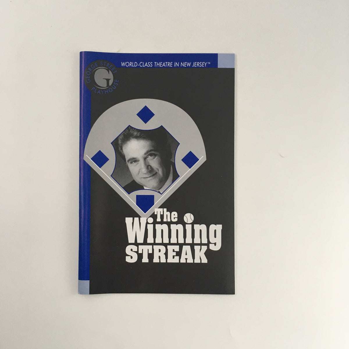 Cover of the play 'The Winning Streak' by Lee Blessing, presented by George Street Playhouse, showcasing a graphic design featuring a silver and blue color scheme with a photo of a male actor.