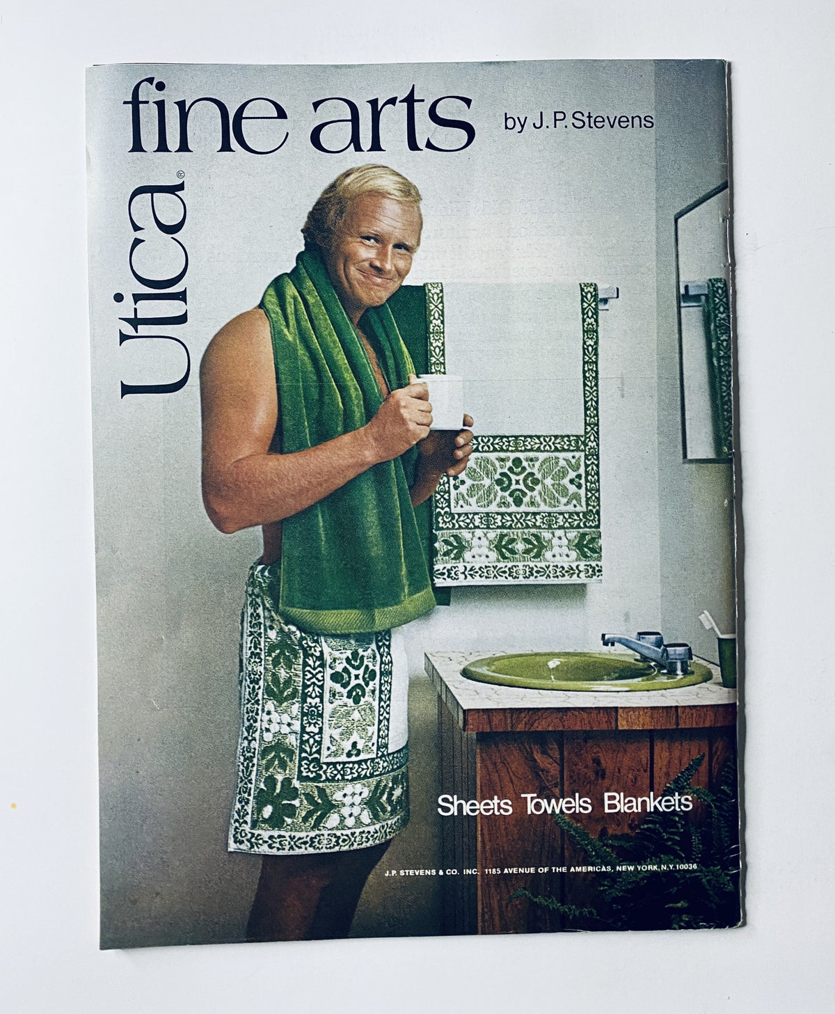 Cover of Utica Fine Arts magazine featuring a man in a green towel and bathrobe holding a cup in a bathroom setting, showcasing products like sheets, towels, and blankets.