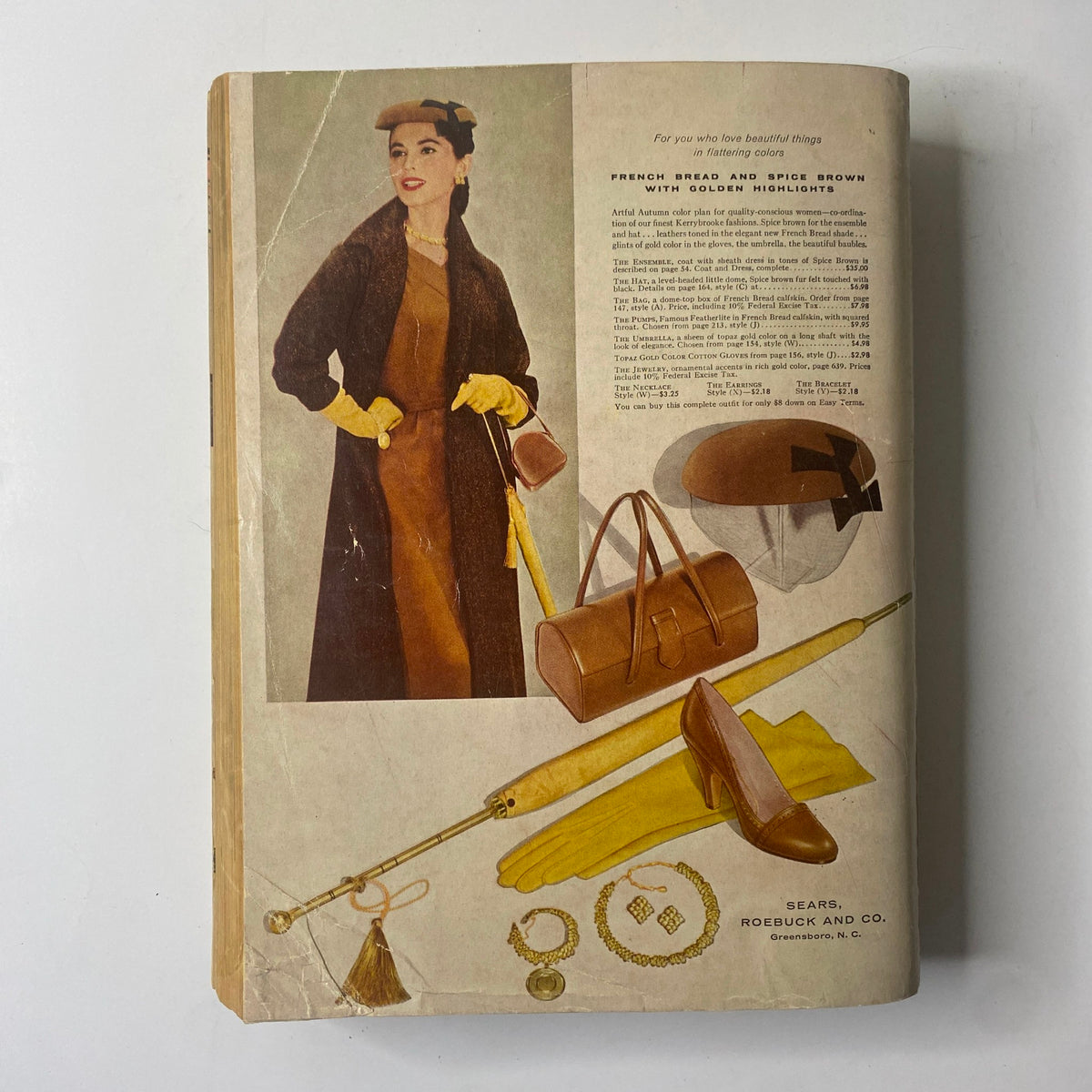 Cover image of the 1956 Sears Roebuck and Co. Fall and Winter Catalog featuring a fashionable woman in a brown dress and coat, accessorized with a yellow hat and gloves, alongside luxury items such as a handbag, umbrella, and footwear, reflecting mid-century fashion elegance.