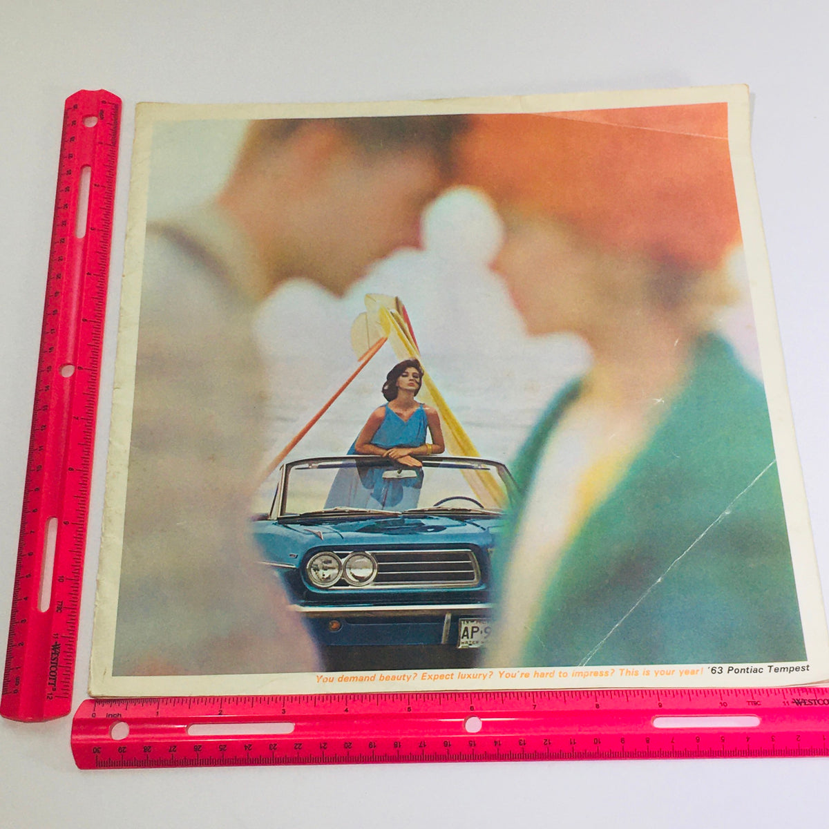 Image of the 1963 Pontiac Tempest dealership brochure against a ruler for scale, showcasing the iconic blue car and a model in a stylish pose.