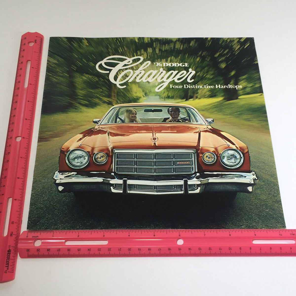 Cover of the 1976 Dodge Charger brochure displayed alongside a ruler for size reference, featuring the text '76 Dodge Charger Four Distinctive Hardtops'.
