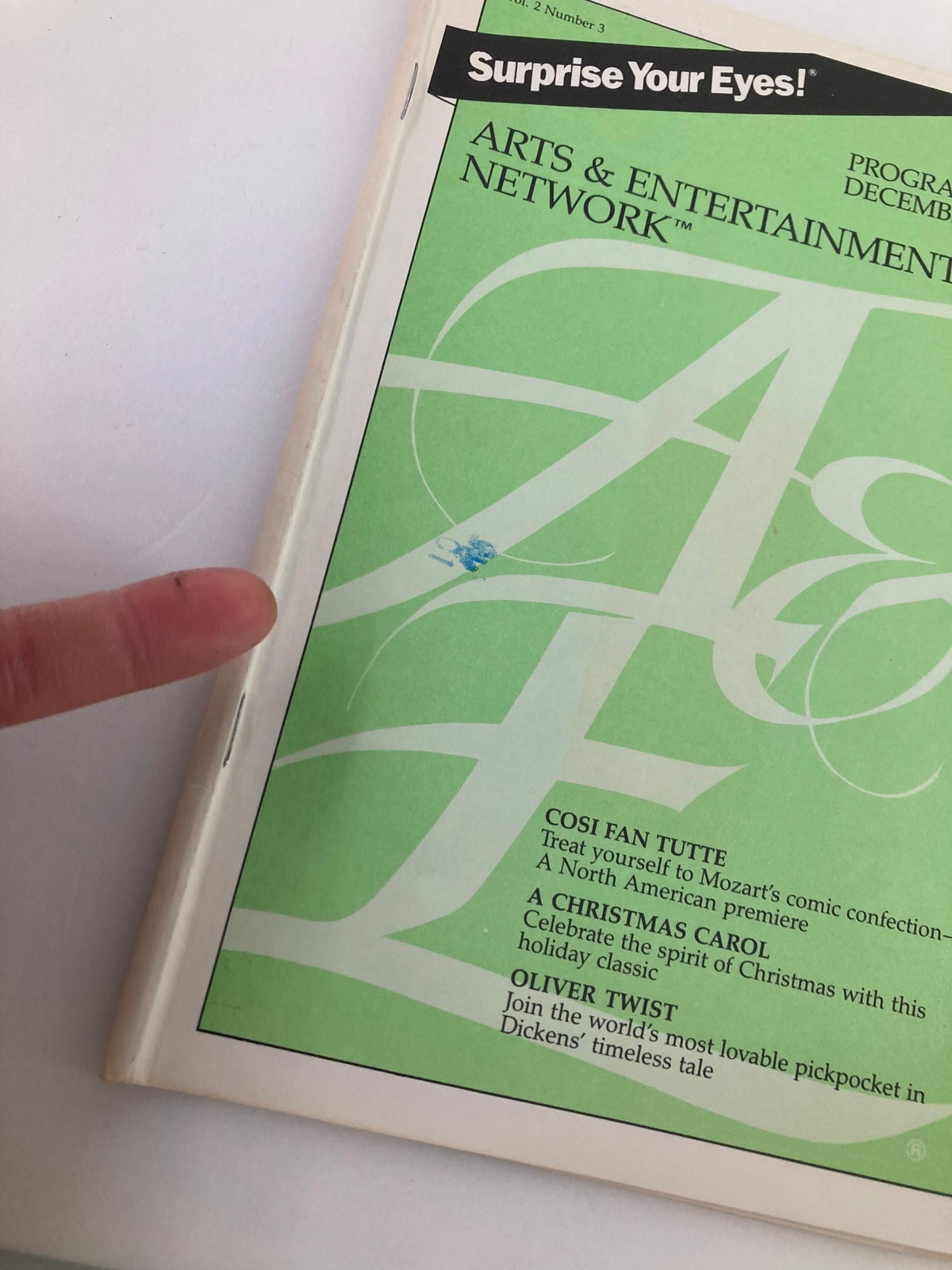 Close-up view of the cover of December 1986 Vol 2 #3 Program Guide from the Arts & Entertainment Cable Network, showing details and highlights like 'Cosi Fan Tutte' and 'A Christmas Carol', and a finger pointing to imperfections.