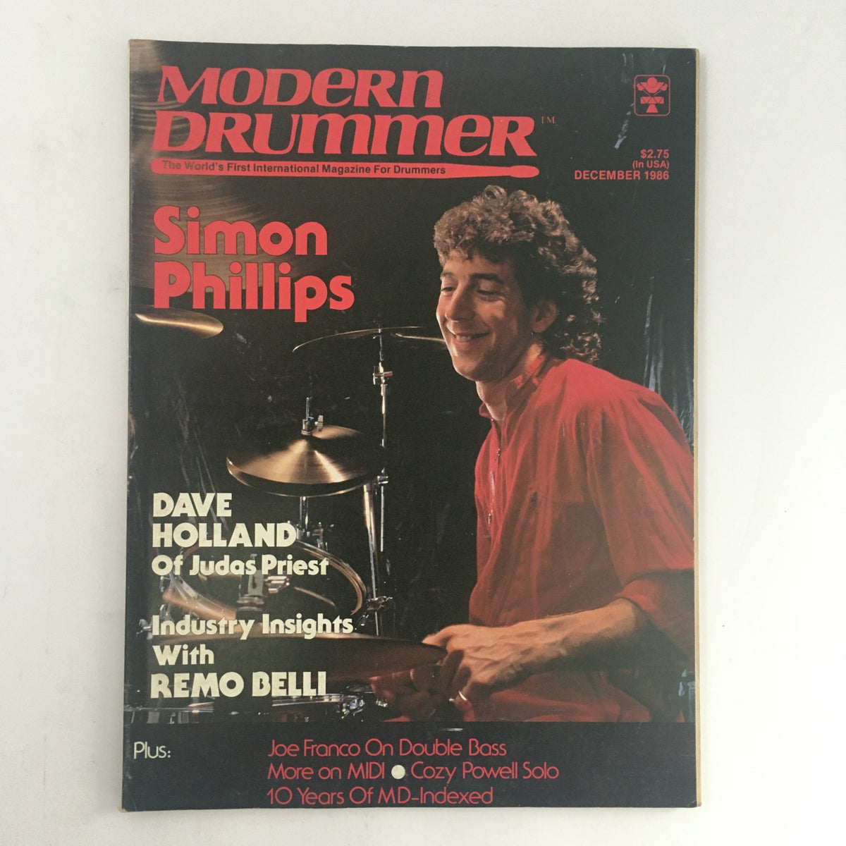 Cover of Modern Drummer Magazine December 1986 featuring Simon Phillips, with a background photograph of him playing the drums, along with text highlighting Dave Holland and industry insights with Remo Belli.