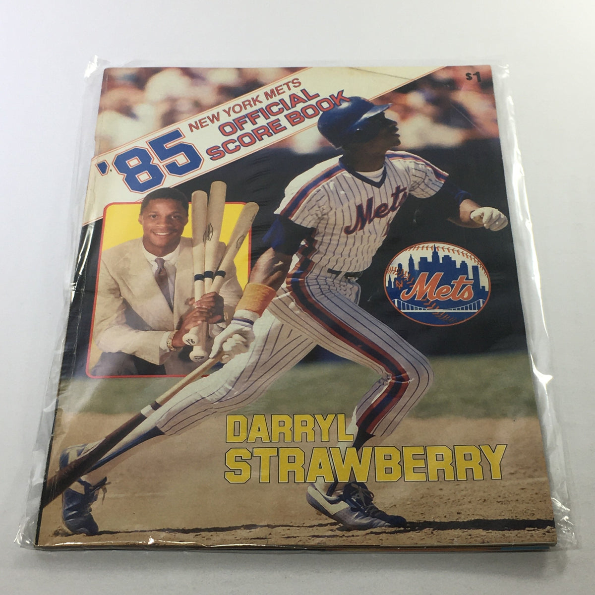 1985 New York Mets Official Score Book featuring Darryl Strawberry, showcasing a vintage design with bright colors, depicting Strawberry in action on the field, and including a photograph of him holding baseball bats. The score book is housed in a plastic protective covering.