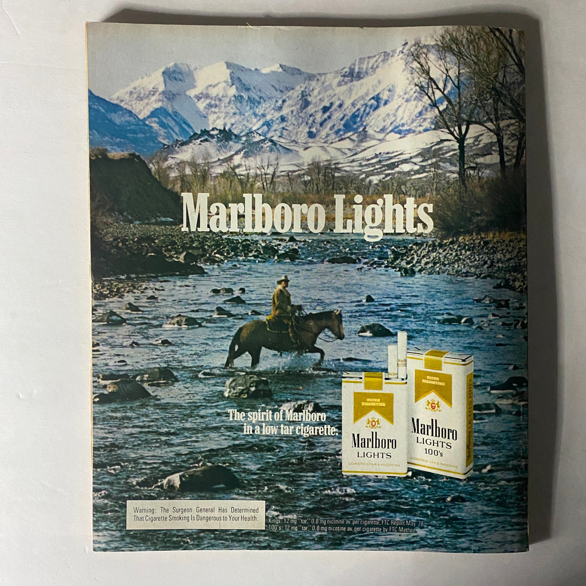 Marlboro Lights cigarette advertisement showcasing a cowboy riding a horse by a river against a mountainous backdrop, with the tagline 'The spirit of Marlboro in a low tar cigarette'. Features the product packaging of Marlboro Lights, emphasizing their low tar content.