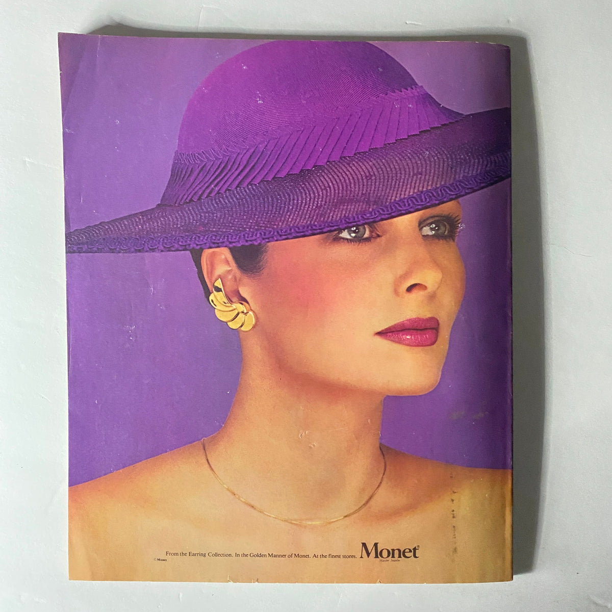 Advertisement for Monet jewelry featuring a model wearing a purple hat and gold earrings, from Harper's Bazaar Magazine February 1980.