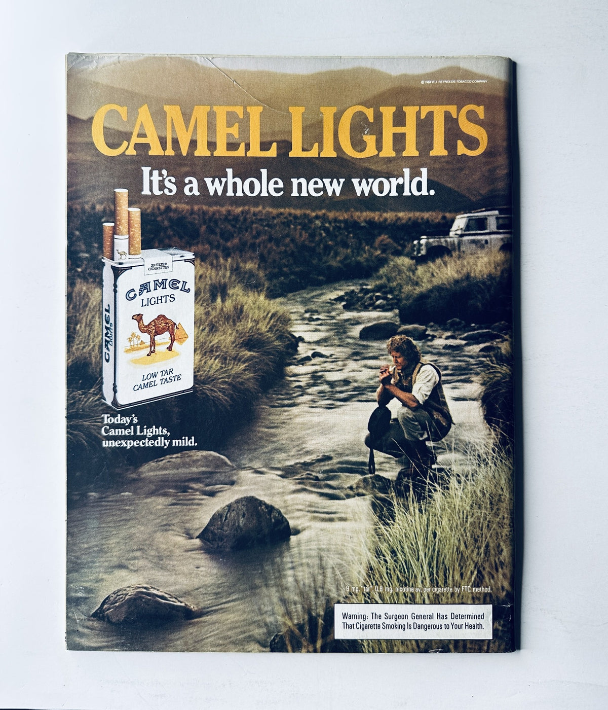 Vintage advertisement for Camel Lights cigarettes, showcasing the slogan 'It's a whole new world' with a scenic background and a man sitting by a stream.