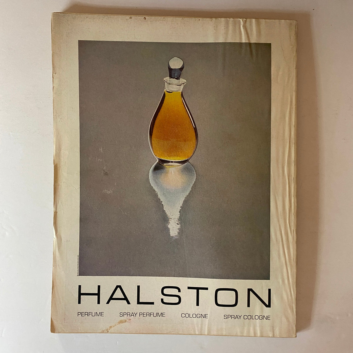 Vogue Magazine April 1976 featuring a Halston perfume advertisement showcasing a striking bottle of yellow perfume, reflective on a smooth surface, accentuating vintage 1970s aesthetics.