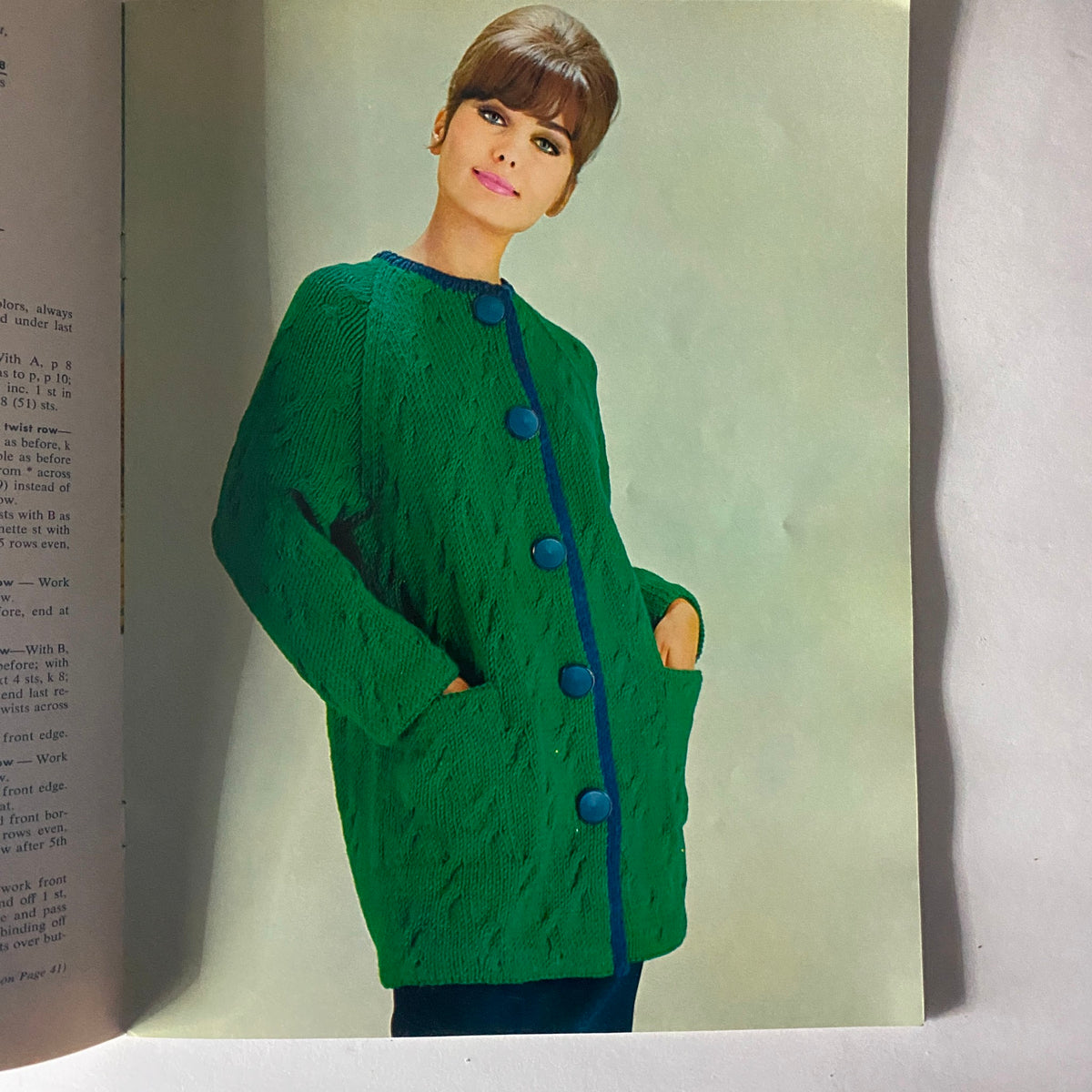 Inside page of '1963 Cable Knits for Men & Women' featuring a model in a vibrant green cable-knit coat with blue buttons, capturing the essence of 1960s fashion.