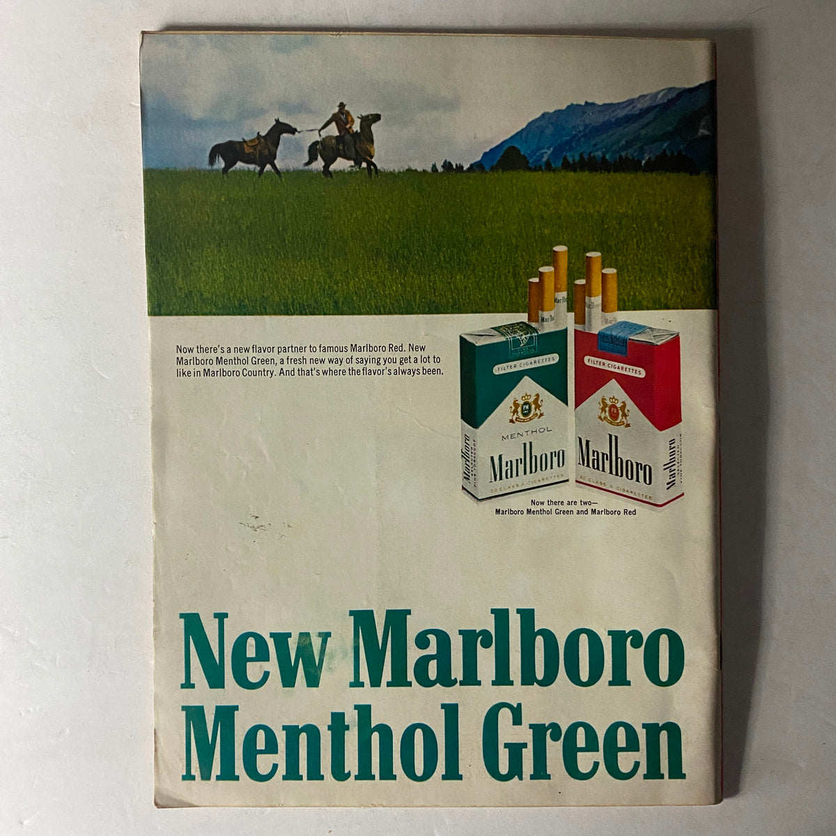 Vintage Marlboro advertisement for Menthol Green cigarettes, showcasing the new flavor alongside Marlboro Red, with a scenic backdrop of cowboys riding horses in a lush field.