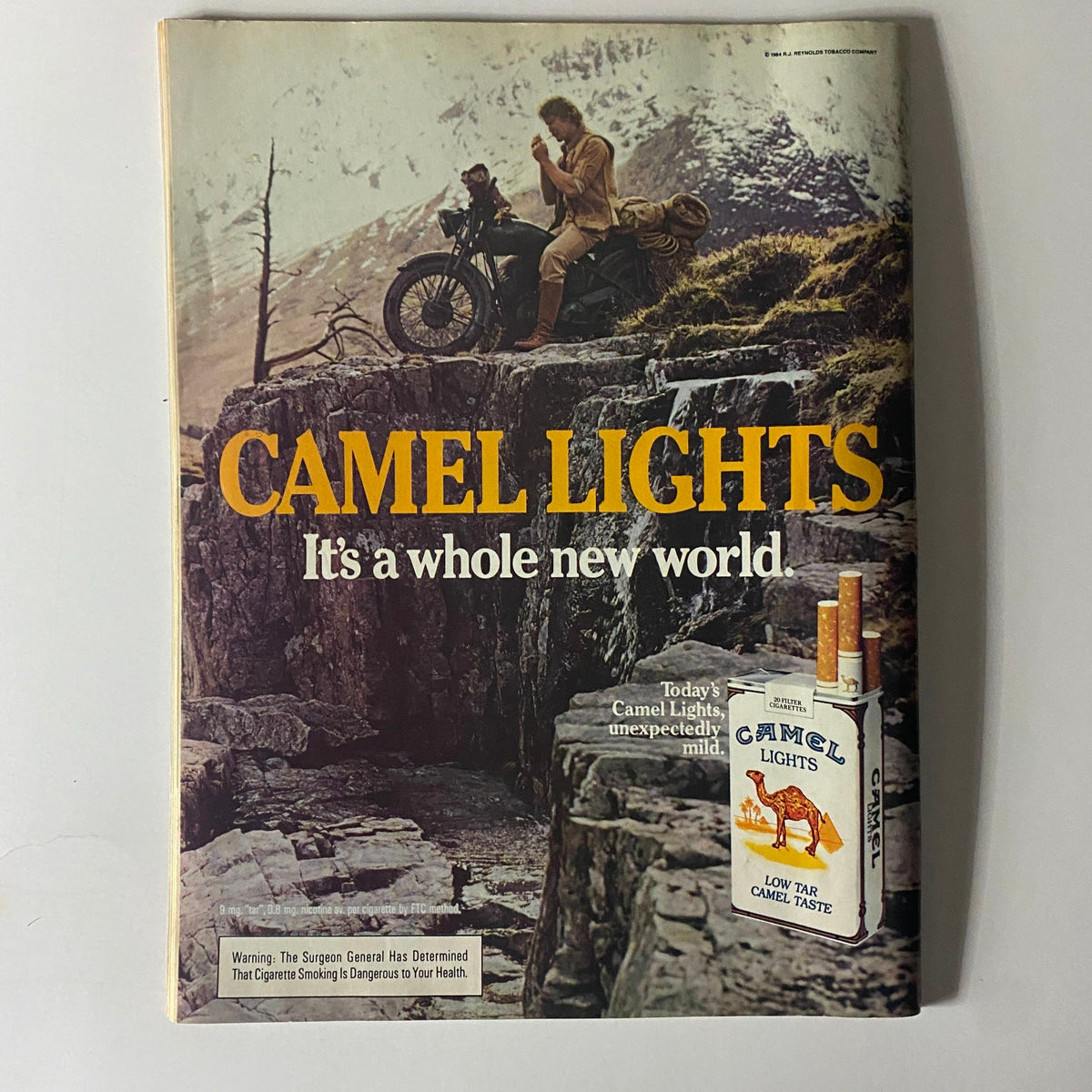 Advertisement from a vintage magazine featuring Camel Lights cigarette, with the slogan 'It's a whole new world' and an image of a man on a motorcycle in a scenic mountainous setting.