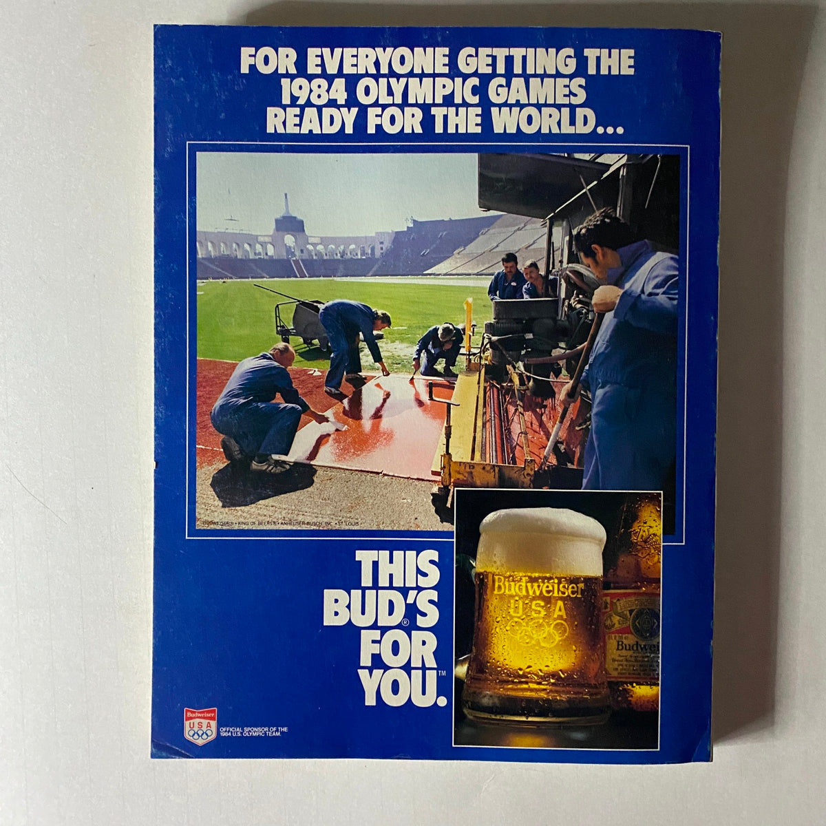 Back cover of the 1984 Official Olympic Guide To Los Angeles Collector's Edition, featuring an advertisement for Budweiser with a photo of workers preparing for the Olympic Games in 1984.