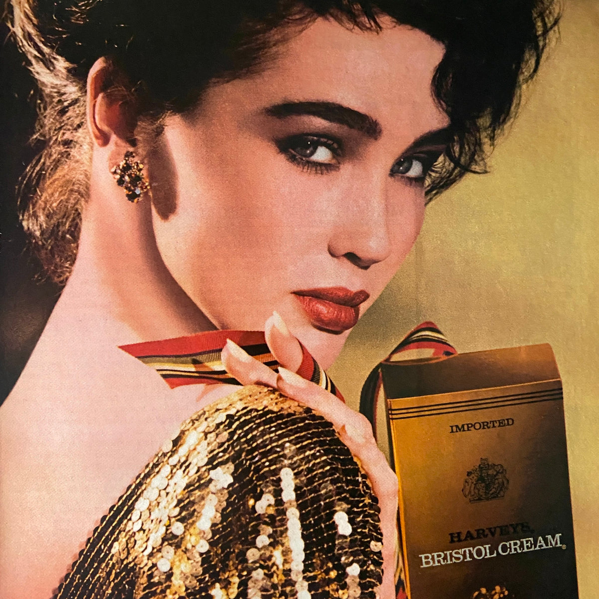 Stylized advertisement featuring a glamorous model holding a box of Harvey's Bristol Cream, set against a sparkling backdrop, emphasizing elegance and sophistication from the Savvy Magazine December 1985 era.