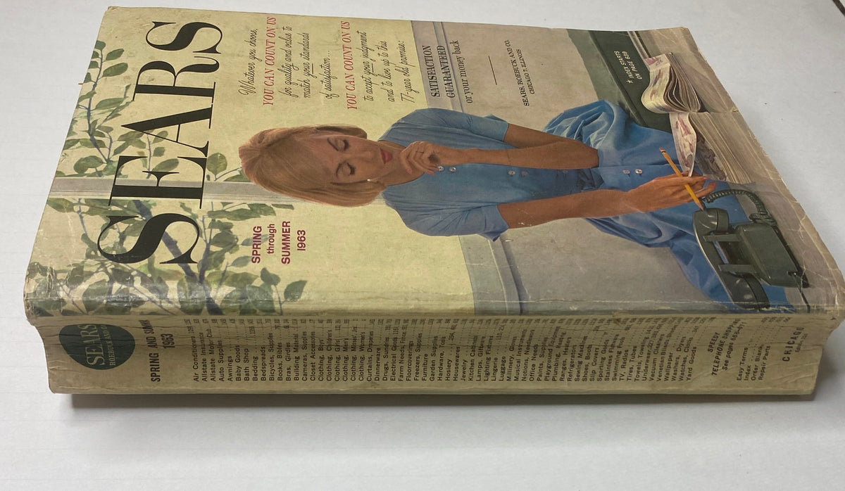Side view of the 1963 Sears Classic Spring and Summer Catalog showcasing its vintage cover design with a woman in a blue dress. The catalog highlights guarantee of satisfaction and features a list of contents on the spine.