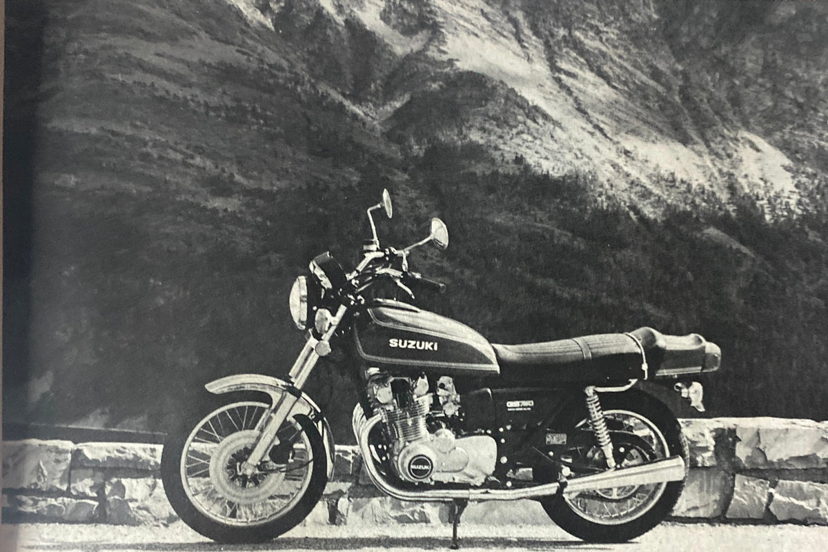 Black and white image of a vintage Suzuki motorcycle against a scenic mountainous background, featured in Cycle World Magazine November 1976.