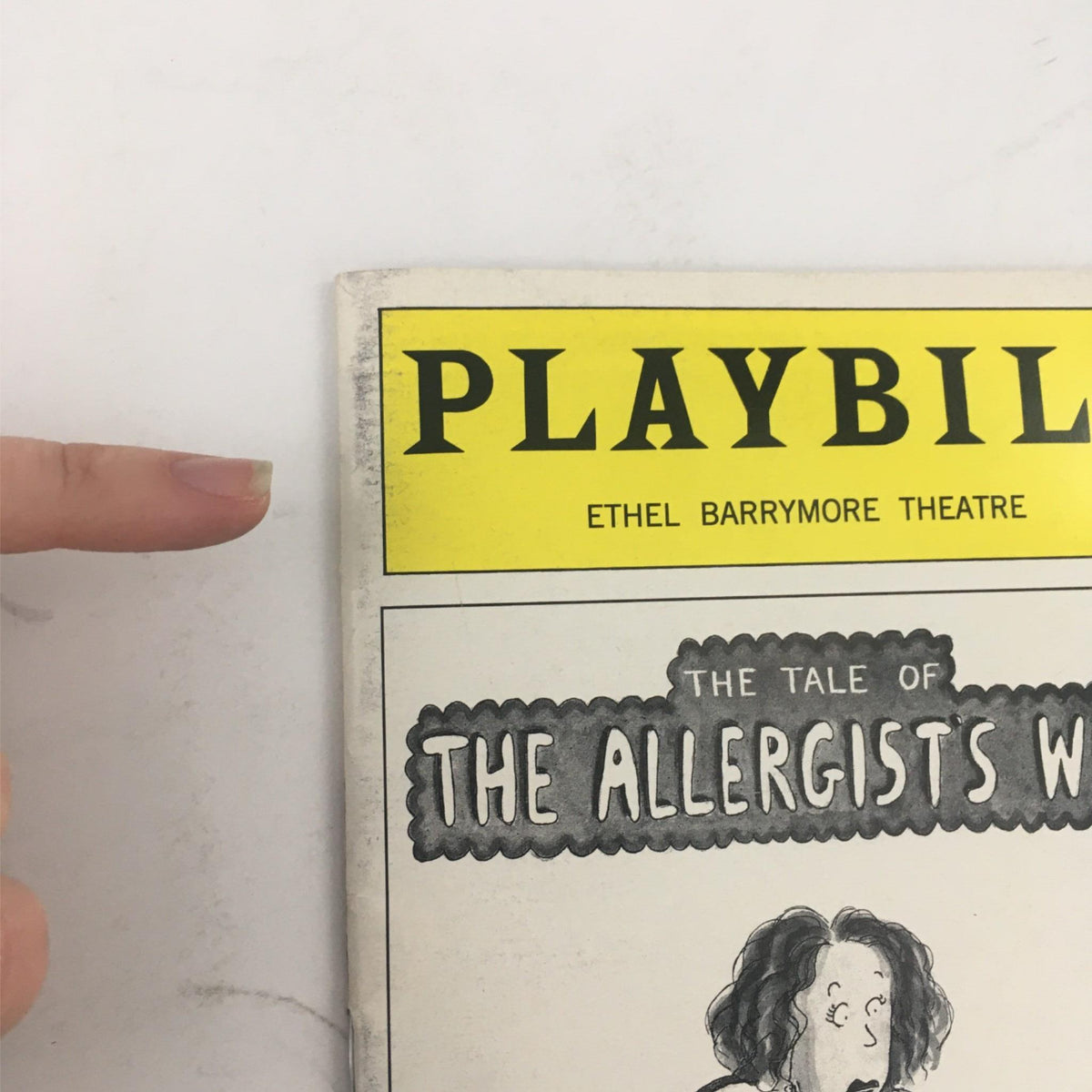 Close-up of the top of the Playbill for 'The Tale Of The Allergists Wife', showing the Ethel Barrymore Theatre logo and a finger pointing to the Playbill title, emphasizing its collectible nature.
