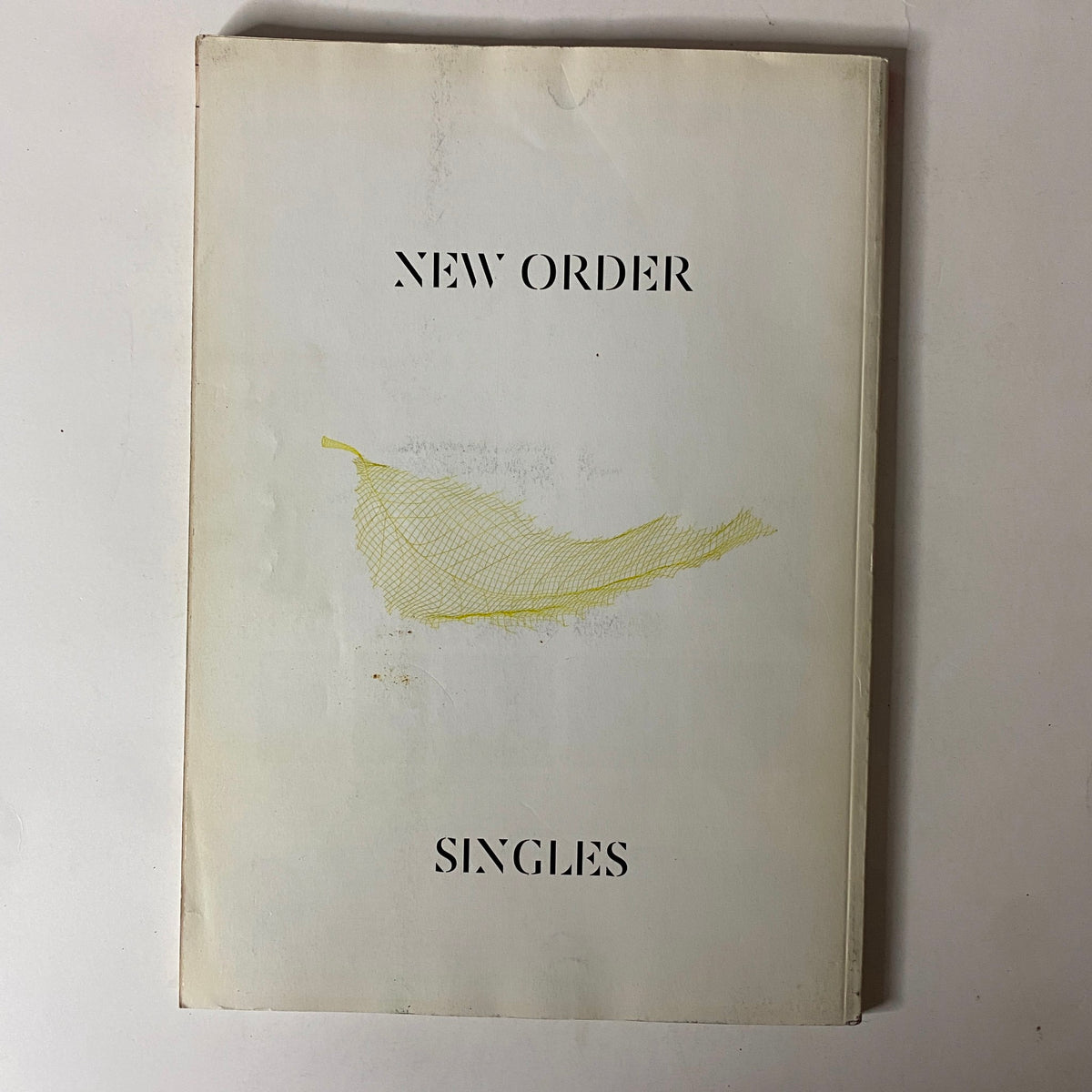 Cover of New Order Singles, a music compilation, showcasing a minimalist design with a simple yellow graphic on a white background.