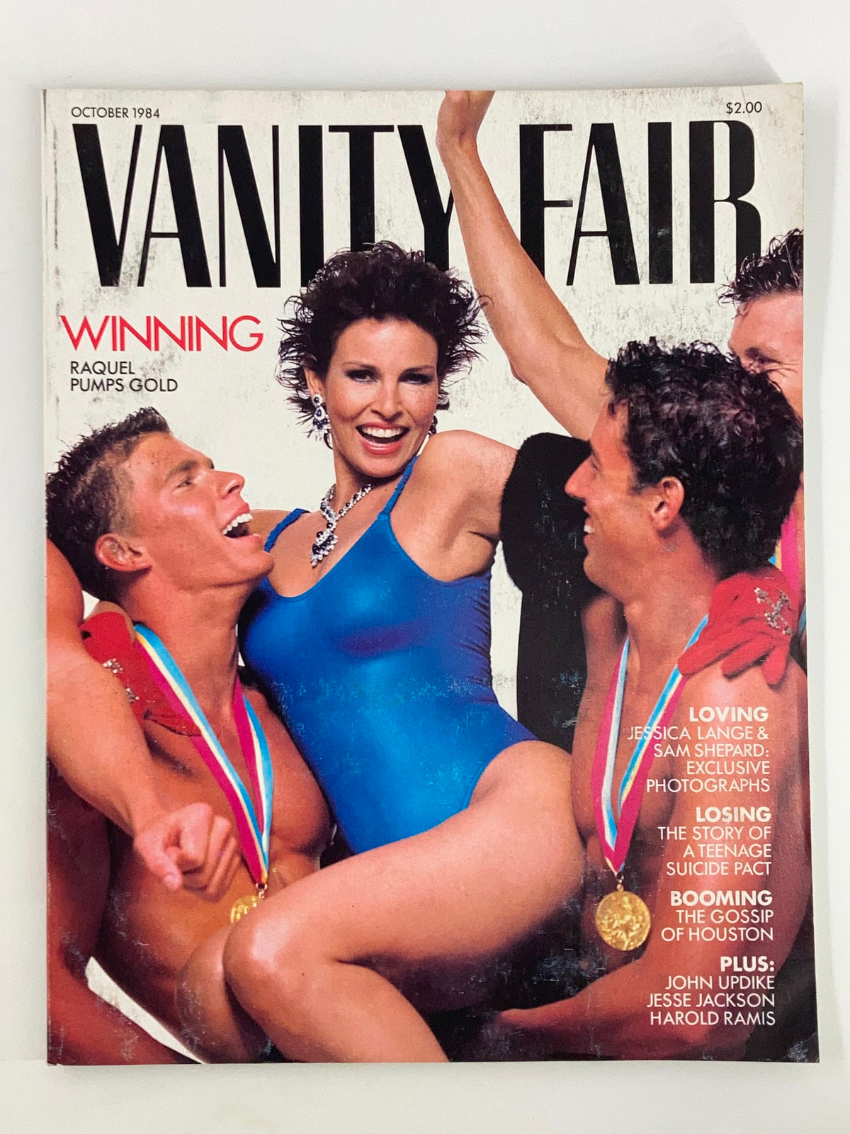 Cover of VTG Vanity Fair Magazine October 1984 featuring Raquel Welch in a blue swimsuit and gold pumps, surrounded by male models, showcasing 80s fashion and glamour.