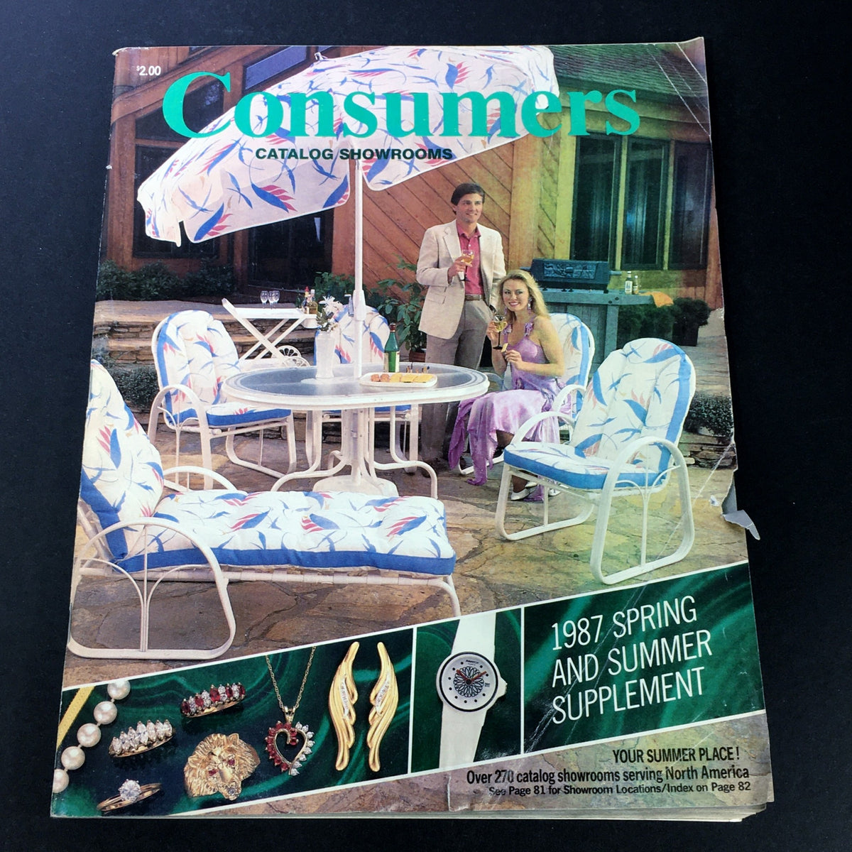 VTG Consumers Catalog Showroom 1987 Spring & Summer Supplement cover featuring a stylish outdoor seating arrangement, two elegantly dressed individuals enjoying drinks, and various decorative accessories showcased along the bottom.