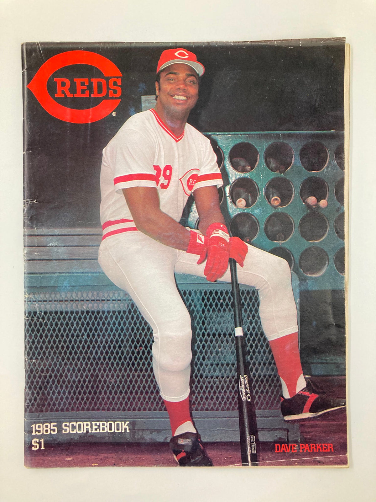 1985 MLB Cincinnati Reds Official Scorebook Program features Dave Parker smiling while sitting with a baseball bat. The scorebook cover includes the REDS logo and indicates it's a collectible item. Good condition with minor wear.