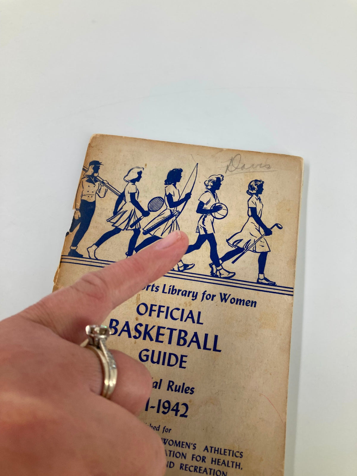 Close-up of the 1941-1942 Official Basketball and Officials Rating Guide for Women and Girls, highlighting the cover design with a finger pointing at the illustrations.