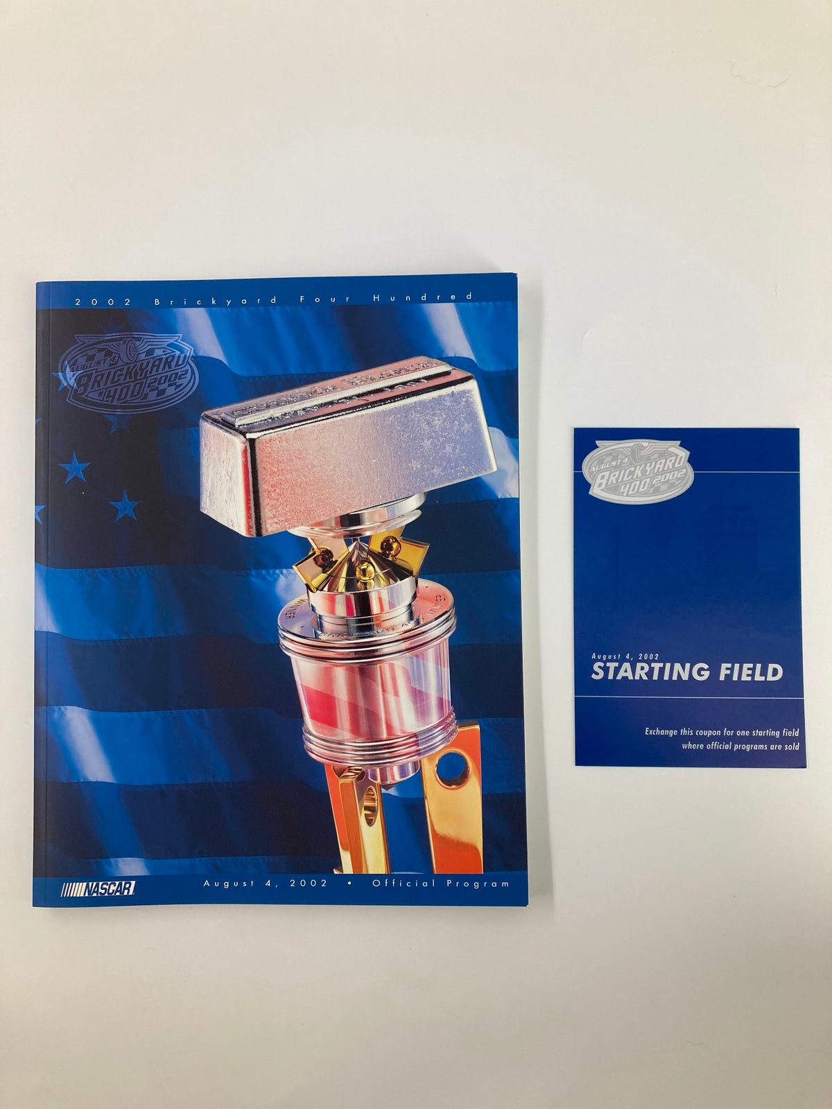 August 4 2002 NASCAR Brick Yard 400 Starting Field Official Program featuring a silver trophy on a blue background along with a coupon exchange card lying beside it.