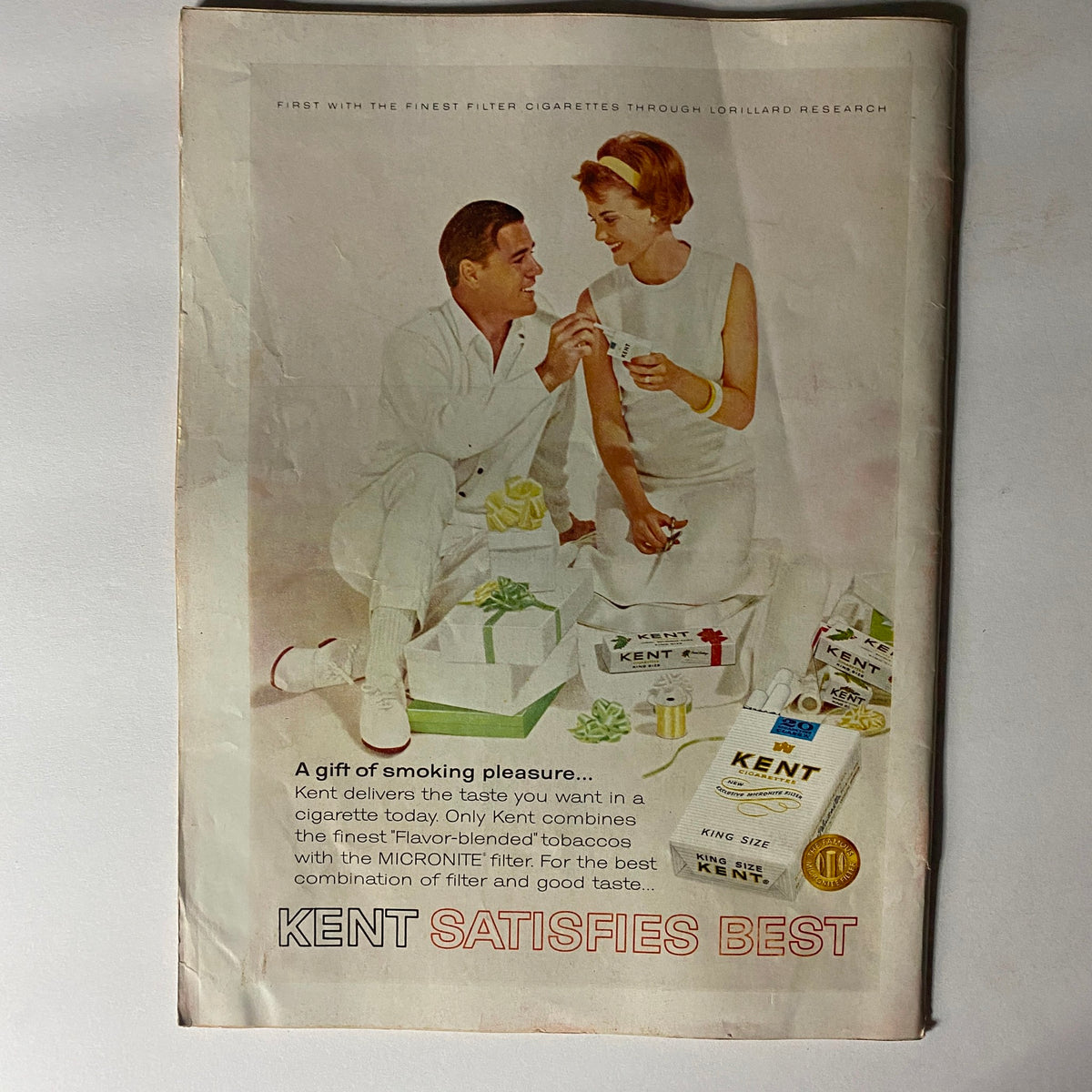 Vintage Kent cigarette advertisement from the 1960s, promoting the brand with a couple exchanging gifts, emphasizing the slogan 'Kent Satisfies Best'.