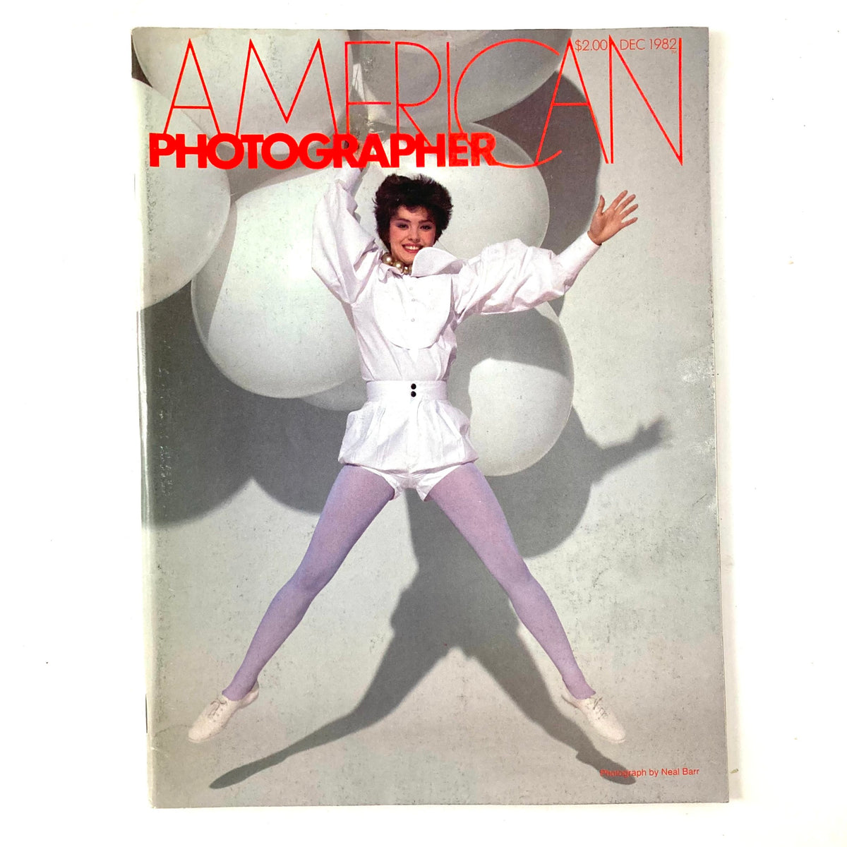 Cover of American Photographer Magazine December 1982, featuring a model in dynamic pose wearing a striking white outfit, with vibrant purple tights and balloons in the background.