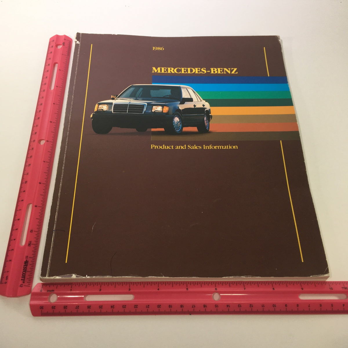 Front cover of the 1986 Mercedes-Benz Product and Sales Information Brochure, showcasing a stylized illustration of a Mercedes-Benz vehicle, with an attractive brown background and colorful stripes.