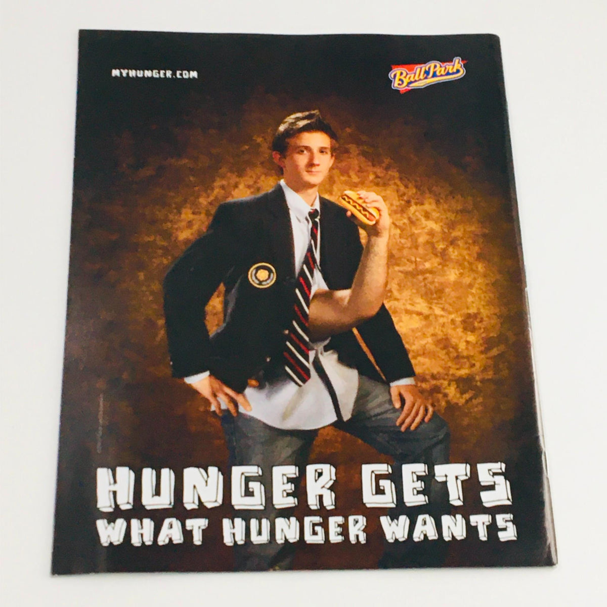 Promotional image for Ball Park featuring a young man in a suit holding a hot dog and displaying a humorous concept with an arm protruding from his stomach, capturing a lighthearted and quirky take on hunger.