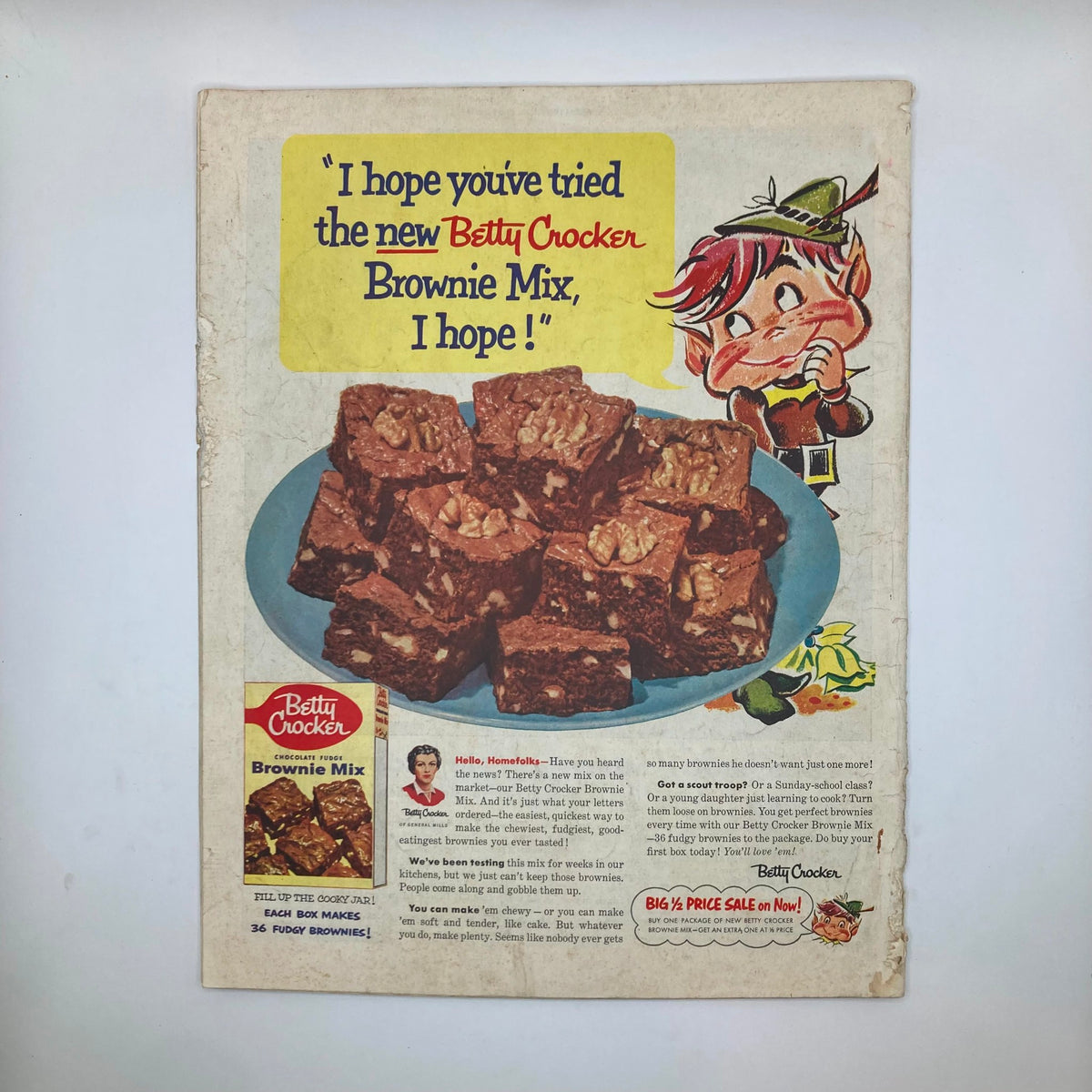 Vintage advertisement for Betty Crocker Brownie Mix showcasing a colorful illustration of brownie squares on a blue plate, with a playful character encouraging consumers to try the new mix, all set against the background of a nostalgic magazine layout. Image from Saturday Evening Post Magazine August 14, 1954.