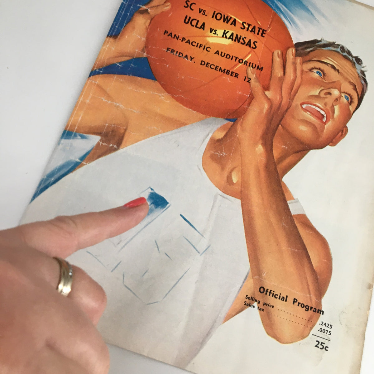 Close-up of the official program from the December 12 1960 NCAA Basketball game between UCLA vs Kansas, highlighting details of the cover and a hand pointing at a specific area.