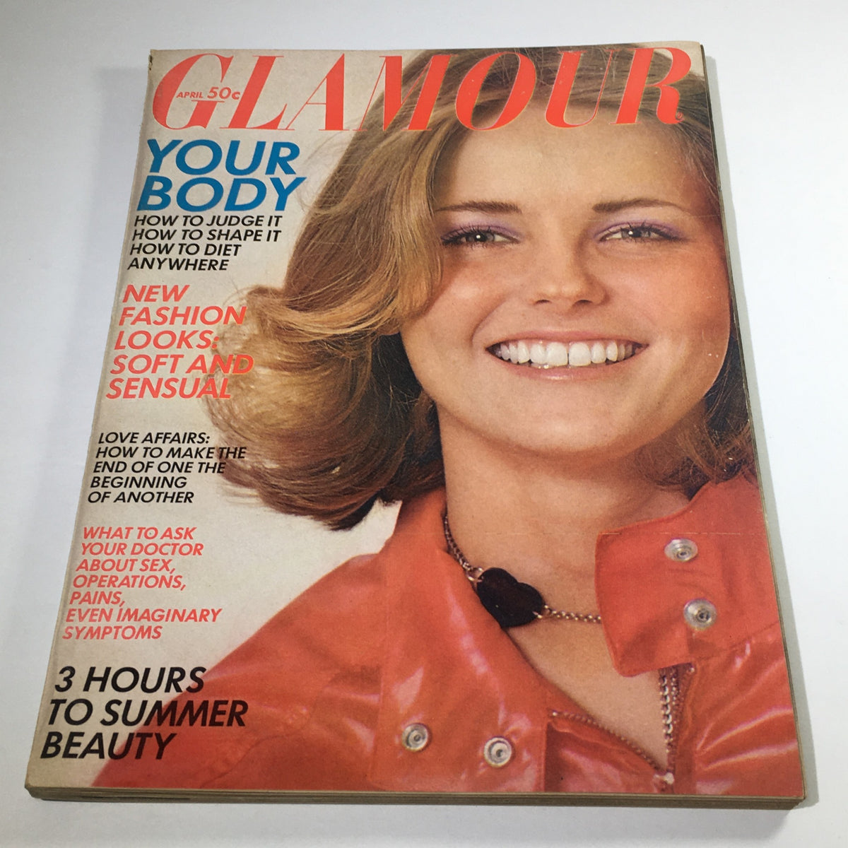 VTG Glamour Magazine: April 1969, featuring a close-up of a model with wavy hair and bright smile, in a vibrant orange jacket, showcasing the cover's bold text and colors with fashion and beauty themes.