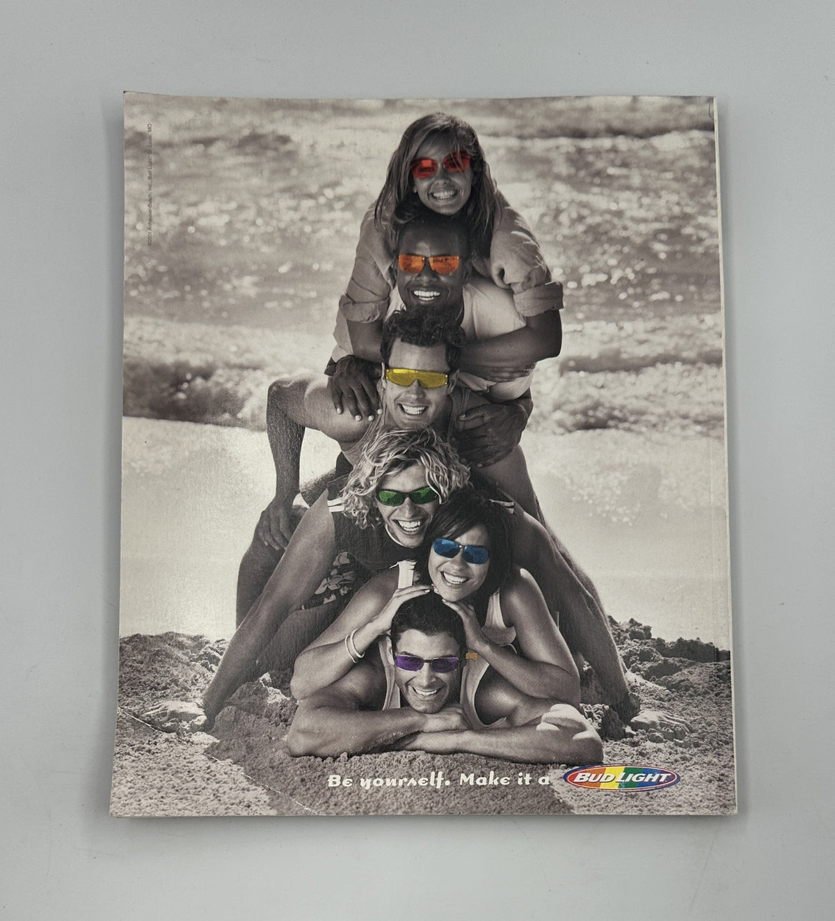 A striking black and white image from Pride Magazine 2001, presenting a group of friends joyfully posing on the beach, wearing colorful sunglasses. The caption reads, 'Be yourself. Make it a Bud Light' promoting inclusivity and fun within the LGBTQ community.