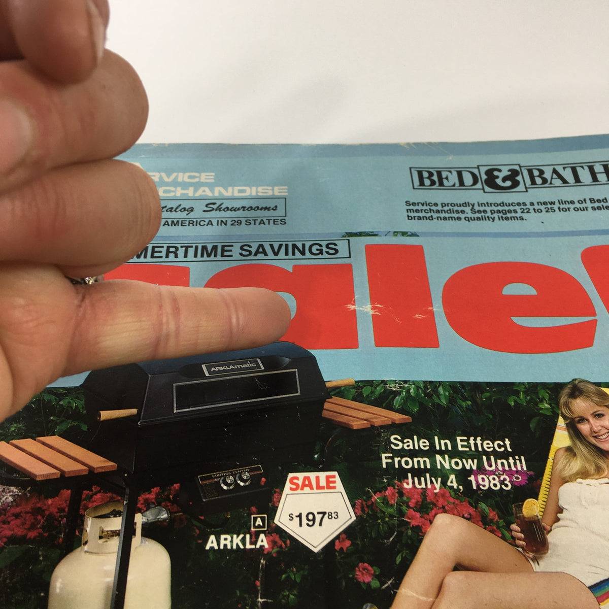 Close-up of the cover of the VTG 1983 Service Merchandise Catalog Showroom Summertime Savings Sale Guide, highlighting the word 'Sale' with a finger pointing at the price of a grill, emphasizing summer promotional offers.